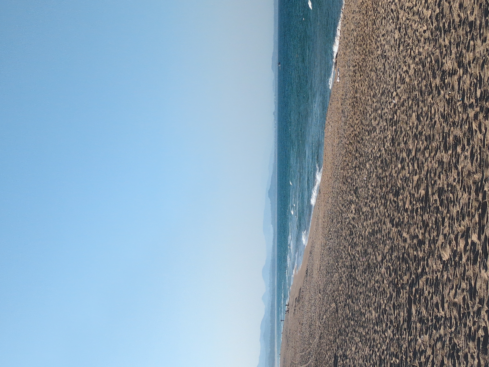The image shows a beach in Tuy Hòa, Vietnam. The scene is divided into three main sections. The left section is a clear, light blue sky. The middle section features the sea, with visible waves and a distant mountain range. The right section consists of a sandy beach with numerous depressions and shadows. Small figures, possibly people, are visible on the beach near the waterline. The scene is well-lit, presumably by daylight.