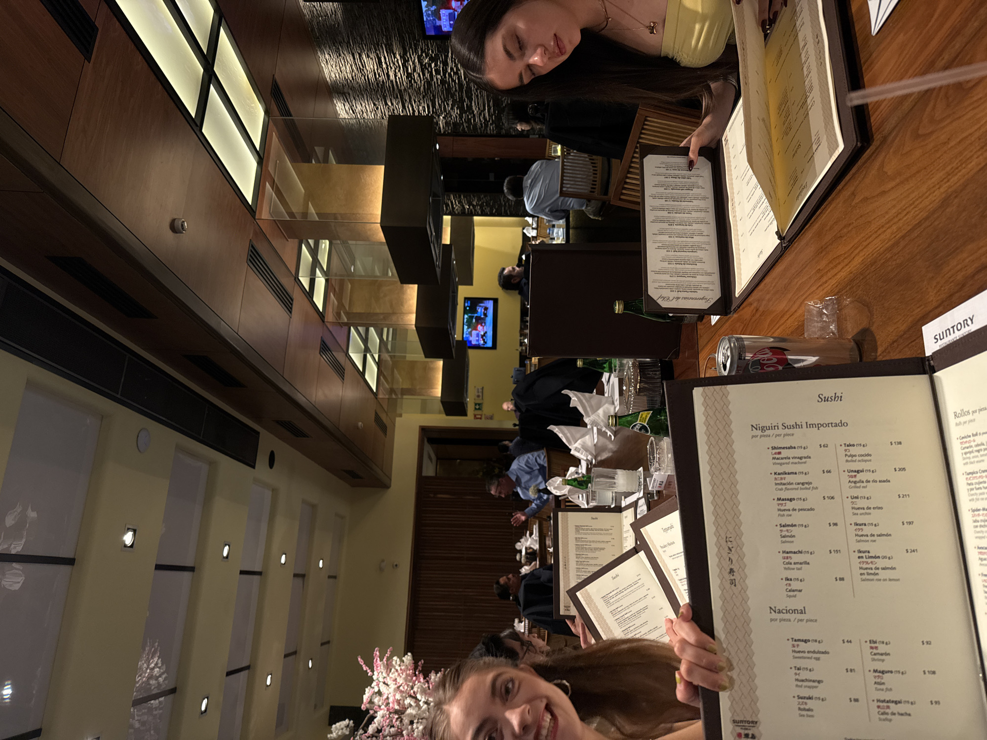 The image appears to be taken inside a well-lit restaurant with a modern interior. There are wooden wall panels, glass dividers, and warm lighting, suggesting a contemporary design. Several people are seated at tables, appearing to dine and interact with each other.
Two women are prominently featured in the foreground. One woman is holding and looking at a restaurant menu, while the second woman, closer to the camera, is smiling and appears to be taking a selfie or being photographed. There are multiple menus on the table, along with drinks and dining utensils, suggesting a meal is in progress.
The restaurant appears to be crowded with other patrons seated at nearby tables. The overall ambiance is upscale and lively.
The image could be taken at Suntory Restaurant in Puebla, Mexico, which is known for its Japanese cuisine. This is based on the "Suntory" brand name on the menu in front of the menu being held, and the type of food on the menu.