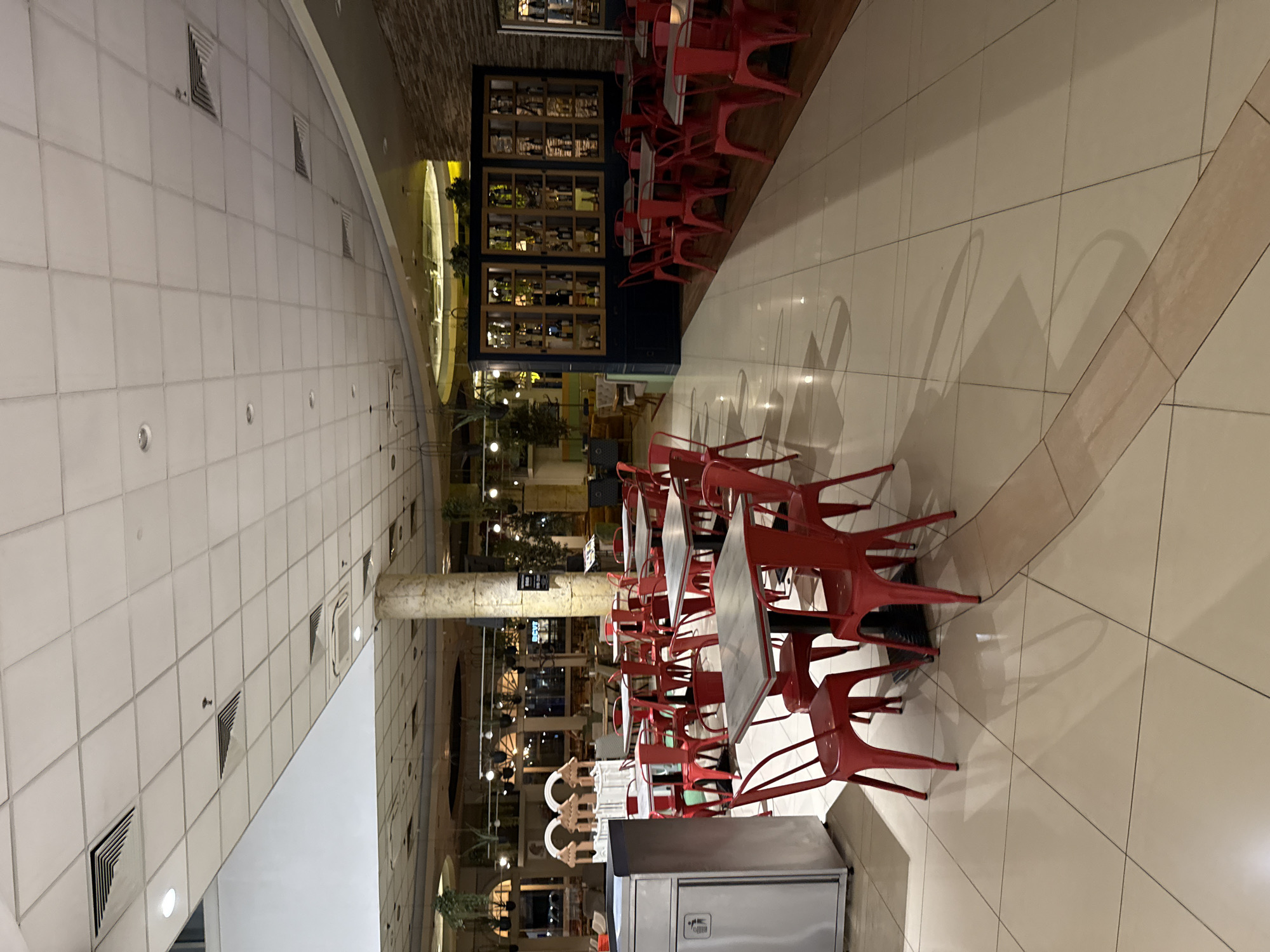 Here is a description of the image:
The image depicts a brightly lit interior space, possibly a food court or a large restaurant. The space is characterized by a high, tiled ceiling with evenly spaced lighting fixtures and ventilation ducts. The floor is covered with large, light-colored tiles with subtle patterns. Numerous red metal chairs are arranged around square tables, suggesting a dining area. A large, cabinet-like structure is visible in the background, filled with bottles and other items, possibly a display or storage area for a restaurant. The overall impression is a modern, clean, and well-maintained environment. There is no visible information to specify the location within the city of Unknown, Unknown.