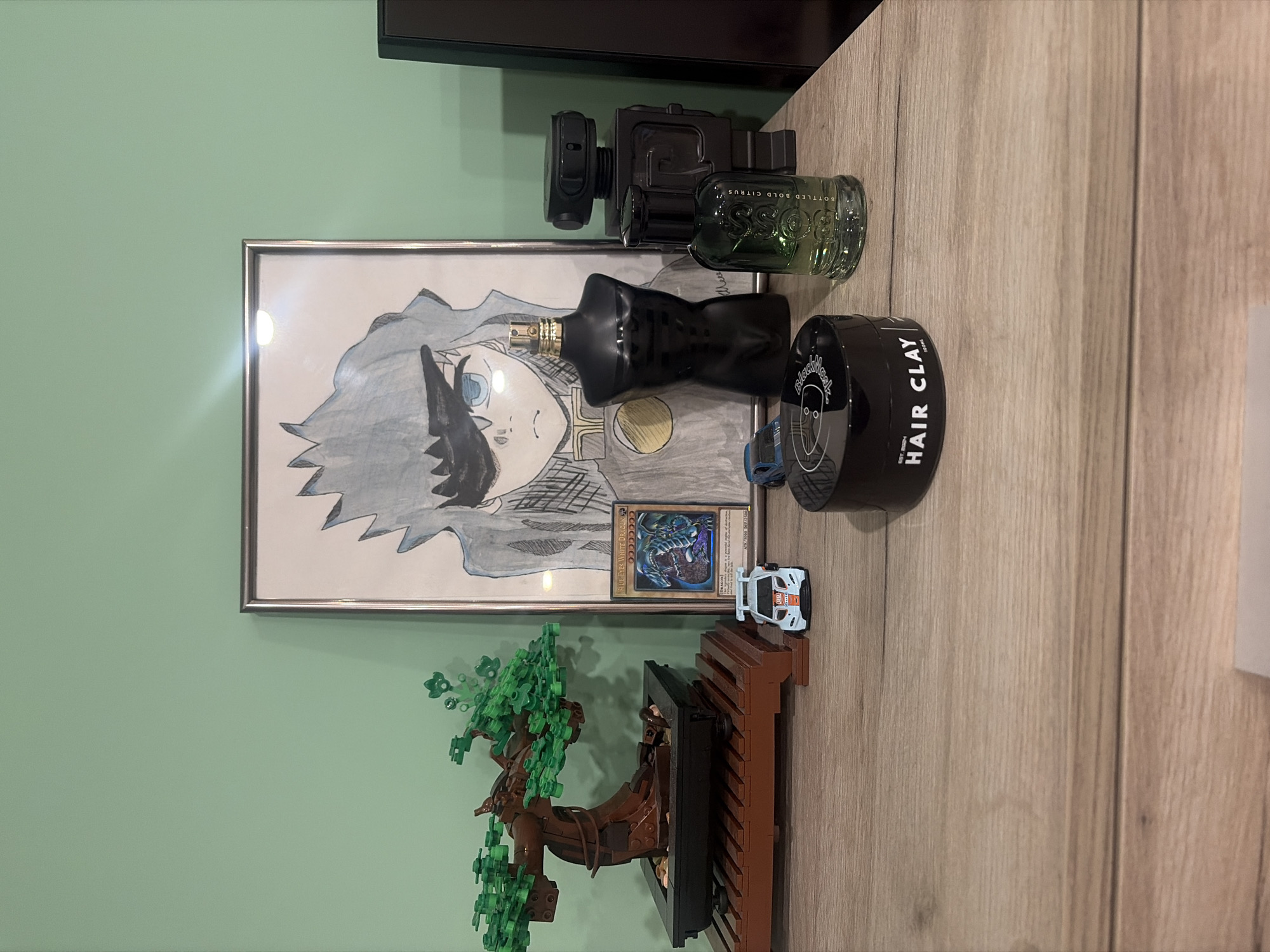 This image depicts a wooden desk surface against a light green wall. On the desk are several objects arranged as decor. To the left, a miniature bonsai tree constructed from LEGO bricks sits on a brown wooden stand. Next to the bonsai, a Yu-Gi-Oh! card featuring "Blue-Eyes White Dragon" is displayed. In front of the card, a white toy race car with orange and blue accents is placed.
Dominating the center of the composition is a framed drawing of an anime-style character with spiky grey hair and blue eyes. To the right of the drawing, a black, torso-shaped perfume bottle stands upright. Next to this, a dark grey, robot-shaped object is positioned. In front of the robot, a green glass perfume bottle labeled "BOTTLED BOLD CITRUS" is visible. A black circular container with "HAIR CLAY" written on it rests on the desk in the foreground. The scene is evenly lit, and no individuals are present. The image does not provide any information about the location in Podgumer, Bulgaria.
