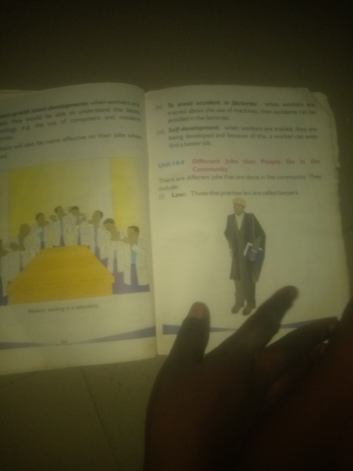 The image shows an open book with text and illustrations. The left page features an illustration of several people in white lab coats standing around a large, yellowish table, with the caption "Workers working in a laboratory." Text on this page discusses workers keeping up with developments and becoming more effective.
The right page contains text that lists points related to avoiding accidents in factories and self-development for workers. A section titled "Unit 14-4 Different Jobs that People Do in the Community" is visible, and under it, the text mentions that people who practice law are called lawyers. Below this text, an illustration depicts a person in formal attire, possibly a lawyer, holding a book and a briefcase.
The image appears to be taken at an angle, with a hand partially obscuring the bottom portion of the book. The overall lighting is dim, suggesting it might be taken indoors. The text and illustrations relate to education and vocational topics. No specific details about the city of Jauro Yinu, Nigeria, are present in the image.