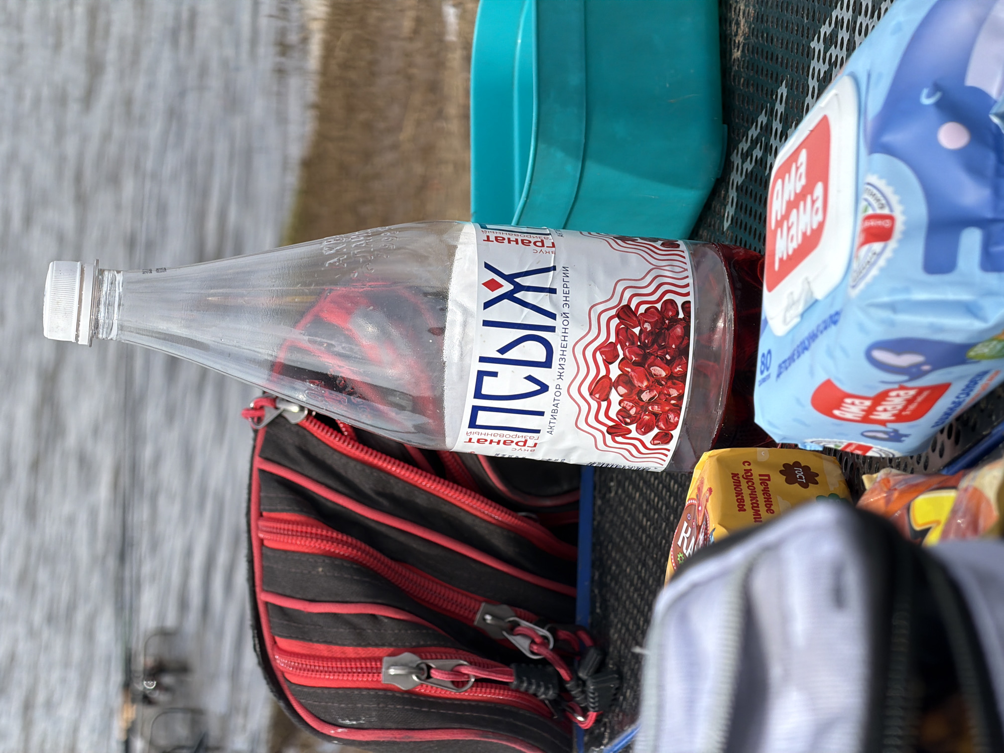 The image captures an outdoor scene by a body of water, likely a river or lake, in Nikolo-Beryozovka, Russia. Various items are laid out on a dark, gridded surface, possibly a pier, boat deck, or a camping table.
In the center, a clear plastic bottle for a carbonated beverage named "ПСЫХ" (Psykh) is prominent. Its label describes it as an "АКТИВАТОР ЖИЗНЕННОЙ ЭНЕРГИИ" (Activator of Vital Energy) with a "гранат" (pomegranate) "вкус" (taste), featuring an illustration of pomegranate seeds. Adjacent to the bottle is a blue package of "Ама мама" (Mama Mama) baby wipes, adorned with an elephant graphic.
Other items include a black and red zippered bag, likely containing personal belongings or fishing tackle, given the faint presence of fishing rods in the blurred background near the water. A yellow package of "Печенье с кусочками Клюквы" (Cookies with cranberry pieces) with a "ГОСТ" (GOST) quality mark is also visible, along with another partially obscured snack package and a grey zippered pouch. A turquoise plastic container or bucket is situated towards the top of the frame.
The background clearly shows the shimmering surface of the water, and the scene appears to take place during daylight hours, suggesting a leisure or fishing outing.