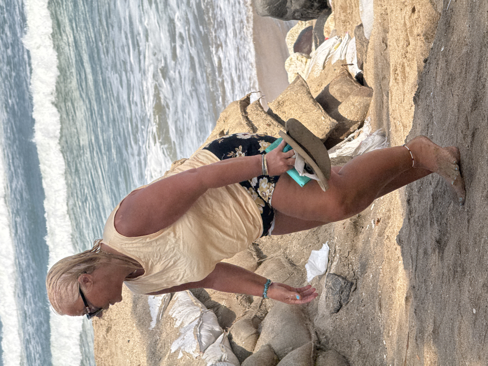 The image depicts a blonde-haired woman with sunglasses on a beach in Kuta, Indonesia. She is wearing a light-colored sleeveless top and floral-patterned shorts. The woman is holding a pair of flip-flops and a light blue object in her right hand. She is walking on the sand, with the ocean and waves visible in the background. Sandbags are piled up to her left, suggesting a possible coastal defense or construction effort. The overall scene captures a moment of leisure or activity on a sunny beach day.