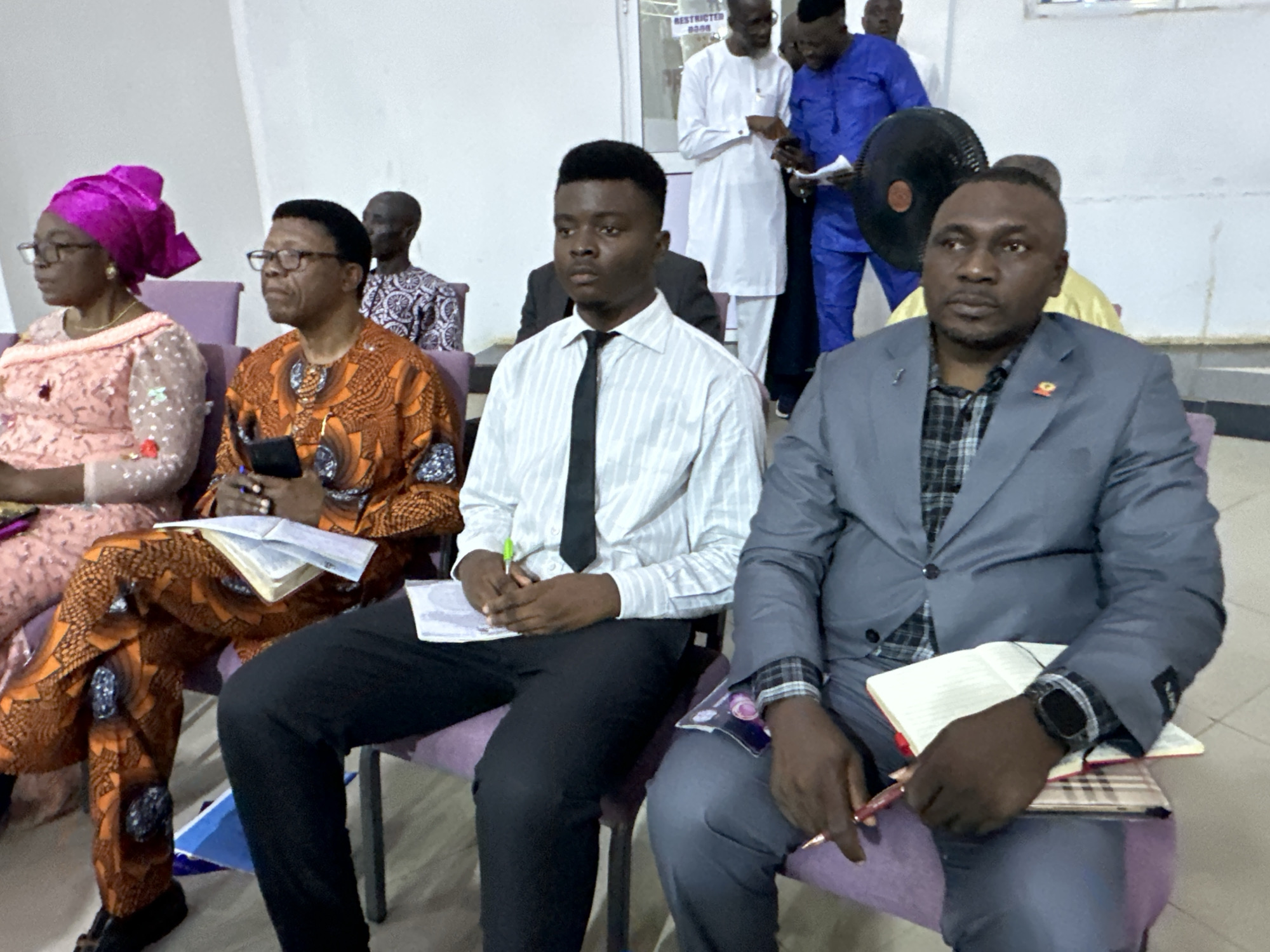 In an indoor setting, potentially a meeting room or place of worship, a group of individuals are seated. In the foreground, a man in a grey suit is looking to his right, holding a pen and resting his left hand on an open notebook. Next to him, a young man in a striped shirt and tie is also looking to his right, holding a pen and a booklet. To the left of these two men, a woman wearing a pink headwrap and a patterned dress is looking forward, with a phone in her hand and a booklet on her lap. Beside her, a man in an orange patterned garment is also looking forward, holding a phone and a booklet. In the background, several other individuals are visible, some standing and some seated, with a large black fan in the middle ground. The room appears to be a typical Nigerian interior, likely within Television, Nigeria, judging by the presence of a sign that reads "RESTRICTED DOOR". The lighting is bright and the overall atmosphere suggests a formal gathering.