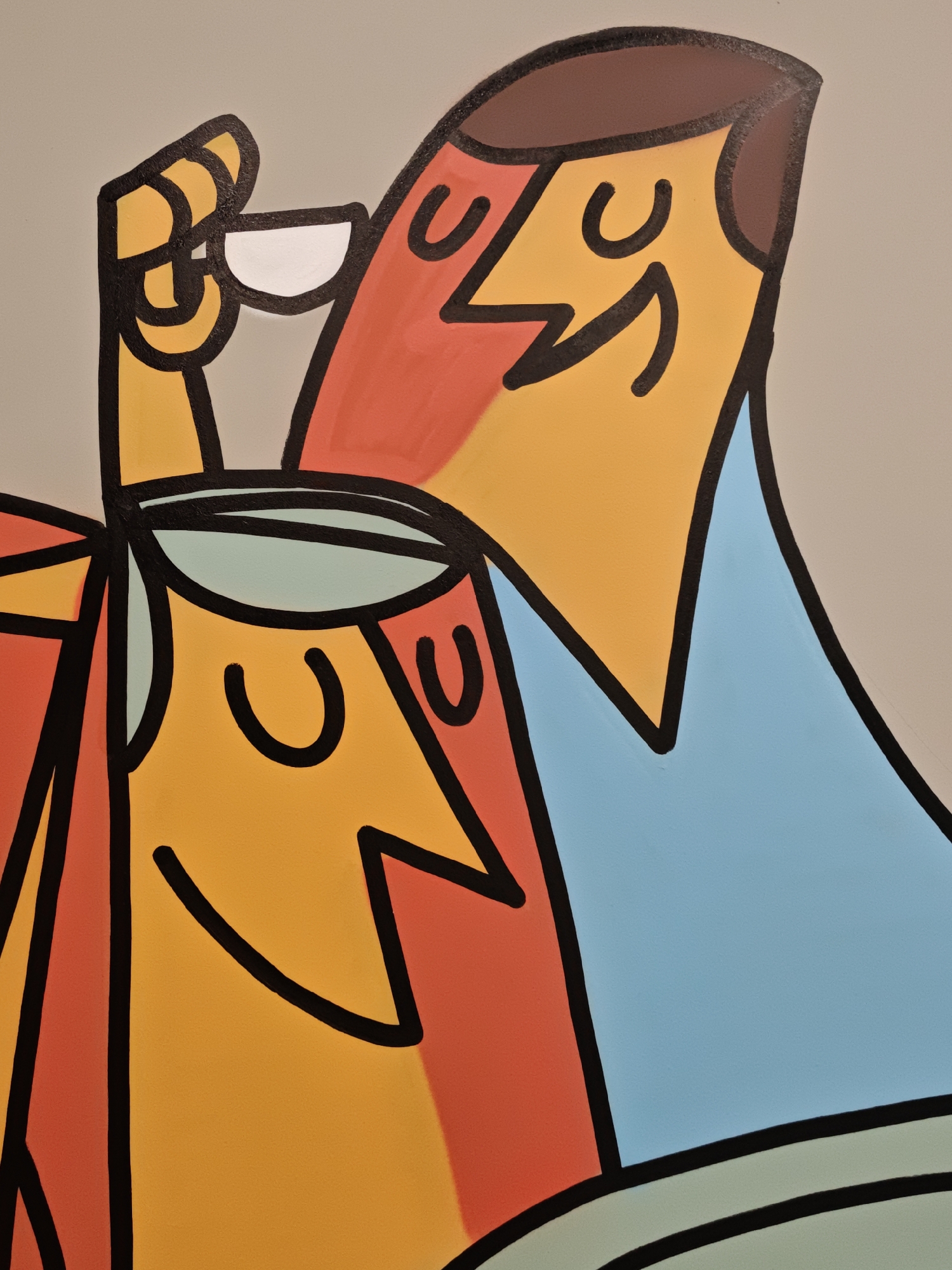 This is a close-up, abstract painting featuring two stylized figures in a light-colored setting, possibly indoors. The figures are depicted with bold black outlines and flat, vibrant colors, including yellow, orange, red, and blue. One figure, in the foreground and to the left, has a light blue oval shape over its head, resembling a cap or headband. Its face is yellow with a black, curved line for a mouth and two U-shaped black lines for eyes. The second figure, in the background and to the right, is taller and also has a yellow face with similar black curved lines for a mouth and eyes. This figure has brown hair and a red and yellow upper body with a blue collar or shirt. One of its hands, colored yellow and outlined in black, is raised to its face, holding a small white object, possibly a cup or a piece of food. The overall style is graphic and simplified, reminiscent of modern art or cartoon illustrations. There is no visible text, indication of time of day, weather, or specific location cues beyond the provided context of Moscow, Russia. The figures appear to be engaged in a shared activity, possibly having a drink or a meal together, and their expressions suggest a calm or pleasant mood.
