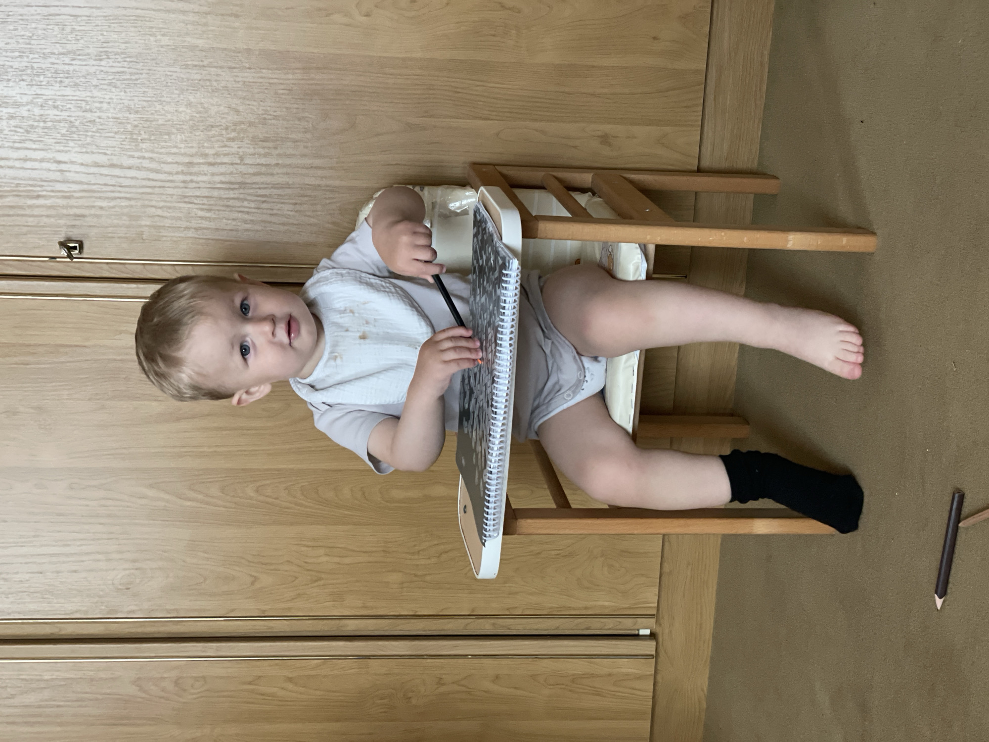 A young child with blonde hair and blue eyes is sitting in a wooden high chair at a small table. The child is wearing a light grey onesie and a bib. They are holding a pencil and looking at a notebook on the table. The child's left leg is bare, while their right leg is covered by a black sock. A wooden pencil is on the carpet in front of the high chair. The background consists of a wooden wardrobe with vertical lines and a wooden door handle. The floor is covered in a brown carpet.