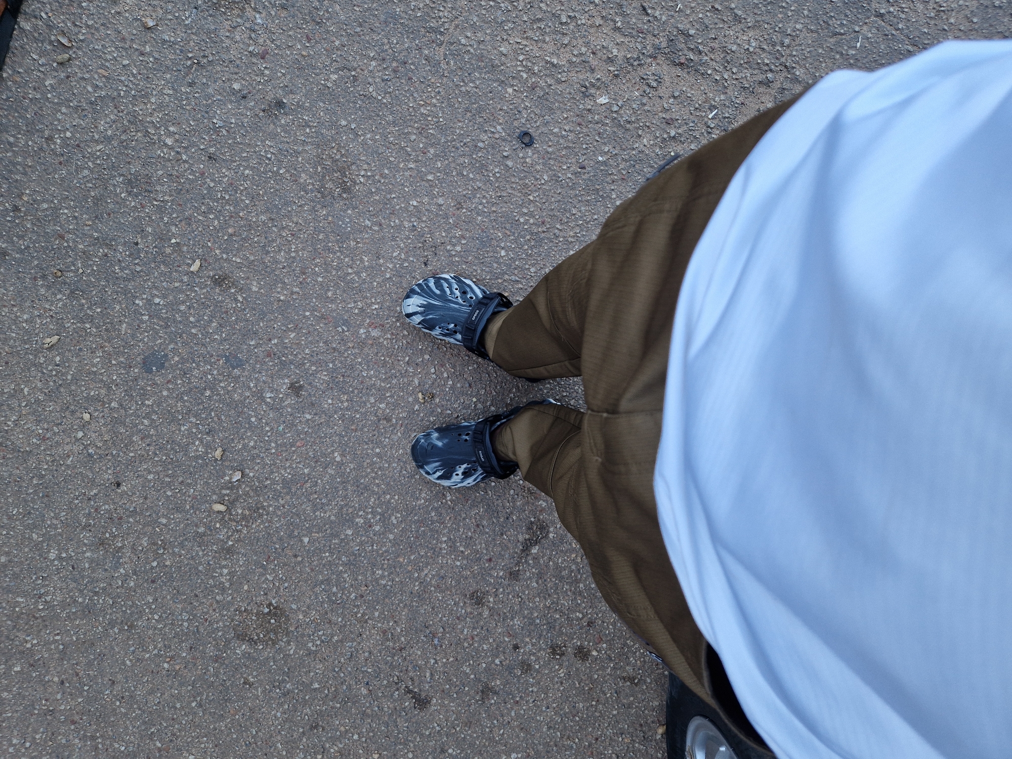 The image is a top-down perspective of a person's legs and feet, standing on an asphalt surface. The person is wearing dark, camouflage-patterned Crocs on both feet and olive-green trousers. A white t-shirt or shirt is visible in the foreground, covering the lower torso. The asphalt appears to be a sidewalk or a road in a city. The lighting suggests it is daytime, possibly overcast. There is no indication of the specific location in Jos, Nigeria, beyond it being an urban outdoor environment.