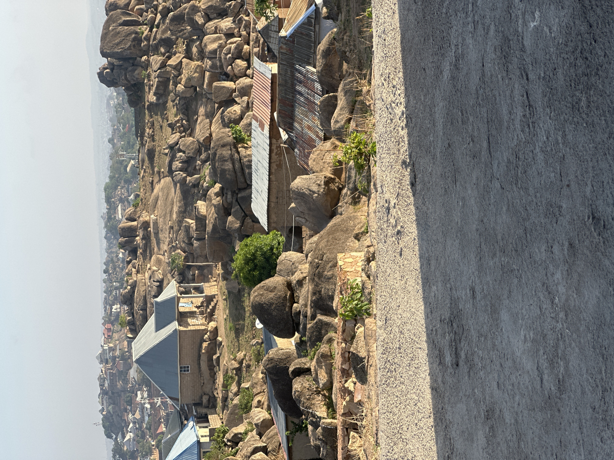 The image shows a landscape in Jos, Nigeria, characterized by a cluster of large boulders and rock formations. Houses are built among these rocks, some with corrugated metal roofs, indicating an adaptation to the terrain. In the distance, there is a hazy view of the city. The foreground shows a section of a gray concrete structure, possibly a road or a retaining wall. The overall environment suggests a rocky, hilly area with a settlement integrated into the natural landscape. There are no visible people or activities depicted.