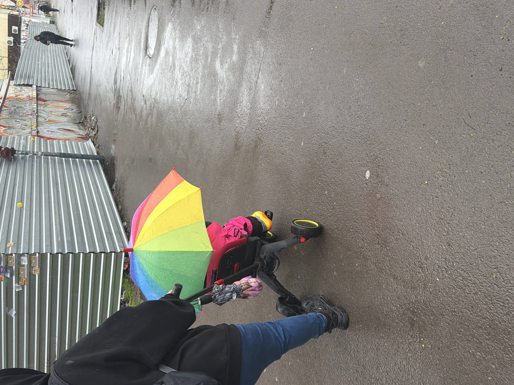 The image shows a person walking in the rain on a wet, asphalt street in what is likely Rostov-on-Don, Russia. The person is pushing a baby stroller covered by a rainbow umbrella. The person is wearing blue jeans, black shoes, and a black jacket. The area to the left has corrugated metal structures and some walls with graffiti. In the distance, another person is walking away from the camera.
