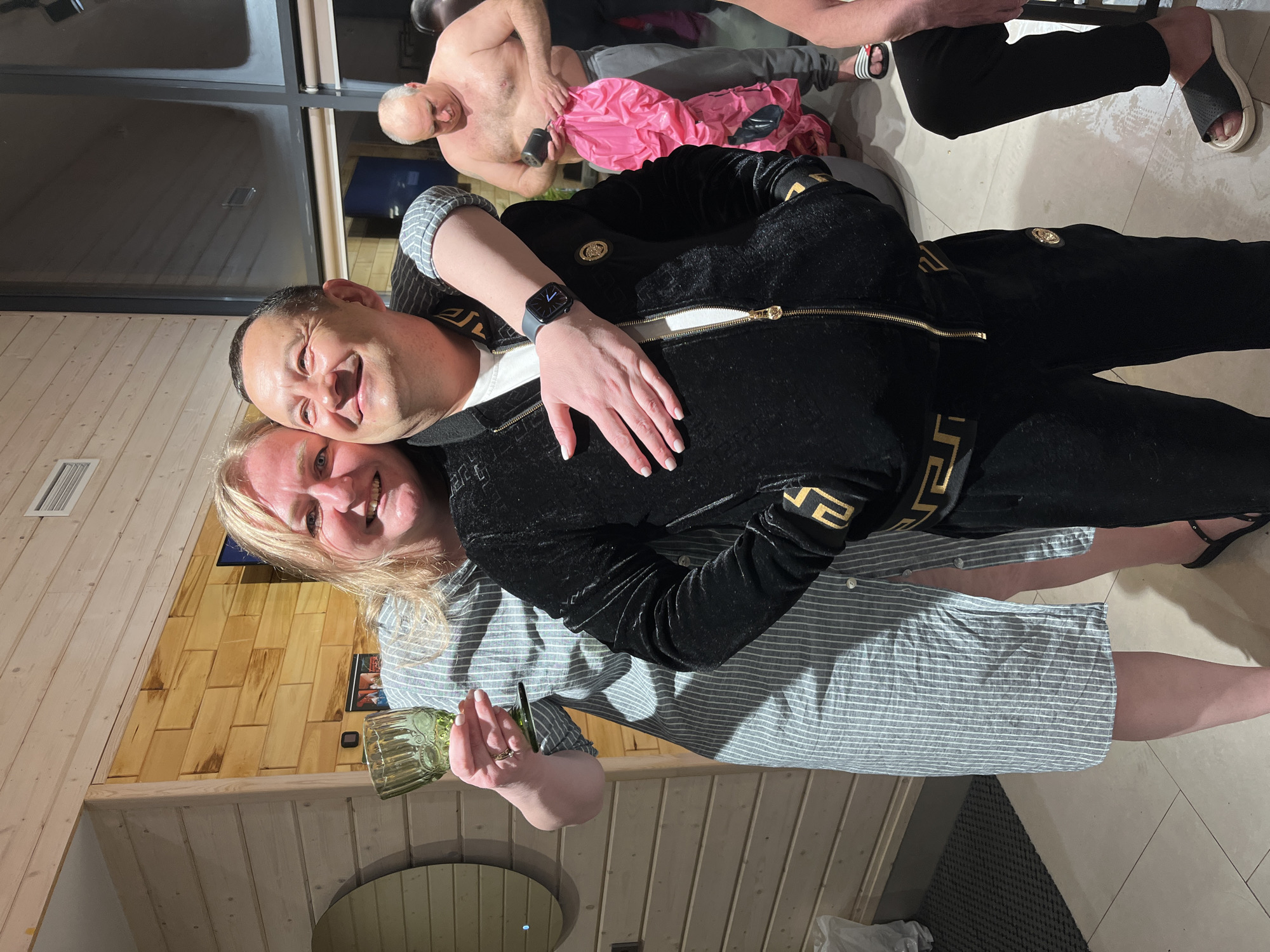 The image depicts a man and a woman posing closely together indoors. The woman is holding a glass, and the man is wearing a black jacket with gold trim. The location appears to be a room with wooden paneling. Other individuals are visible in the background, including a shirtless man. The setting is likely a social gathering or party. There is no explicit evidence to confirm the location as Izhevsk, Russia.