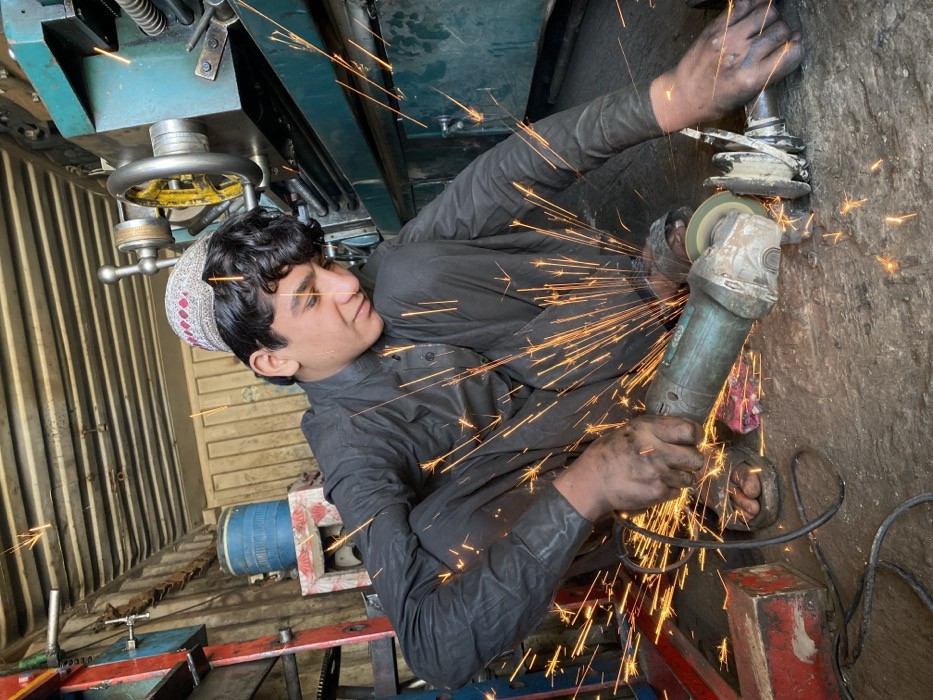 A young male, appearing to be in his late teens or early twenties, crouches on a rough concrete floor within a metalworking shop in Kandahar, Afghanistan. He wears a dark grey long-sleeved tunic and pants, along with a light-colored traditional cap. His hands are visibly soiled. He is actively operating a turquoise-bodied angle grinder, applying its spinning abrasive disc to a metallic, irregularly shaped component, possibly a vehicle part. A dense shower of bright orange sparks emanates from the point of contact, scattering across the foreground. In the background, a large teal-colored lathe machine with prominent handwheels (one yellow, one silver) is visible. To the left, a blue barrel rests on a painted wooden stand, positioned against a wall constructed of corrugated metal sheets. The environment suggests an industrial or repair workshop setting.