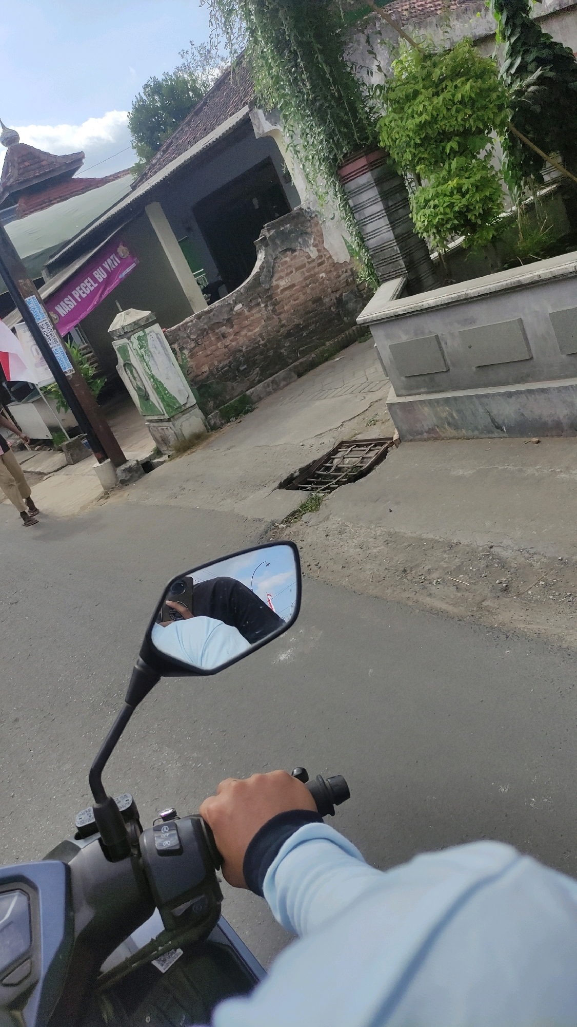 The image is a first-person perspective of someone riding a motorcycle on a street in Wonokromo, Indonesia. In the foreground, a hand in a light blue long-sleeved shirt grips the motorcycle's handlebars. The motorcycle's rearview mirror reflects the rider's head, wearing a dark head covering and holding a smartphone.
The street is paved and appears to have some cracks and a grated drain opening on the right side. To the left of the street, there is a building with a tiled roof and a sign that reads "NASI PECEL BU YANI". The building is adorned with green vines hanging down from above. Adjacent to the building is a weathered brick wall. Further to the left, a person in tan pants and a dark shirt is walking away from the camera. The sky is partly cloudy. The overall environment suggests a residential or commercial area in a tropical climate.