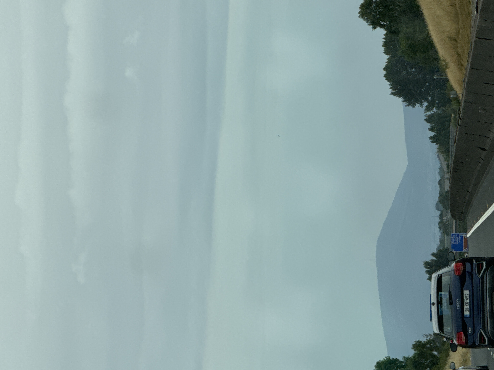 The image captures a road scene, likely a highway, in or near Pill Belan, Ireland. The sky is overcast with varying layers of clouds, creating a muted, atmospheric light. In the foreground, the back of a dark-colored Audi SUV is visible, displaying the license plate "211-KK-492". The vehicle is in motion, suggesting it is traveling on the road.
Beyond the car, the highway stretches out, bordered by concrete barriers on the right side and a grassy embankment with scattered trees on the left. In the mid-ground, a large, hazy mountain range dominates the horizon, its slopes appearing soft and indistinct due to the distance and atmospheric conditions. The vegetation consists of mature trees with dark green foliage and areas of dry, golden grass. There are also indications of other vehicles and roadside signage, but they are not clearly defined. The overall impression is one of a journey through a landscape under a cloudy sky.
