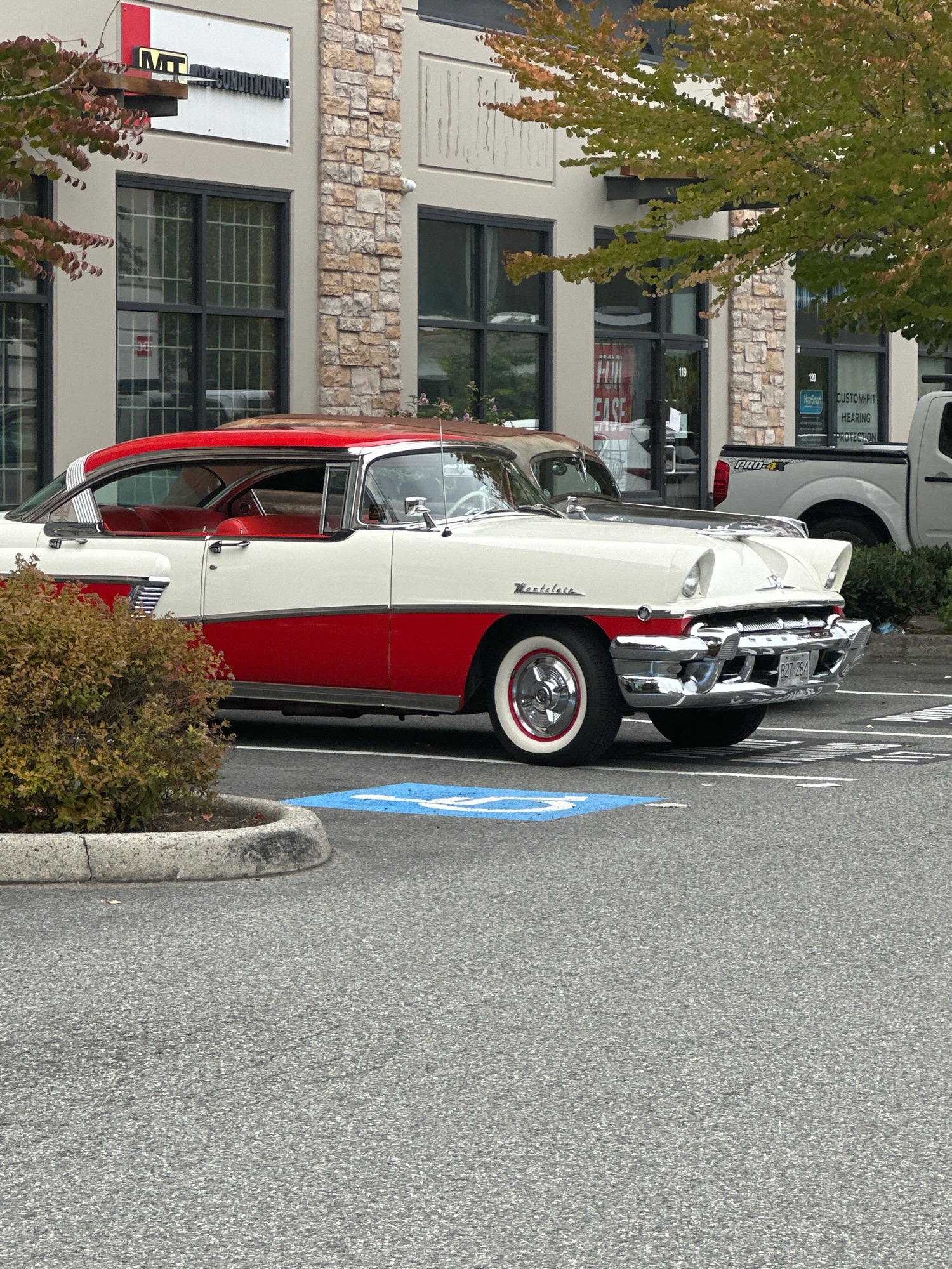 An eye-level medium shot captures a vintage, two-tone red and white Mercury Montclair parked in a lot with a building in the background. The car is angled towards the right of the frame, showcasing its chrome grille and accents. A blue handicapped parking symbol is visible on the asphalt in front of the car. The building behind the car features large windows, brick detailing, and signs indicating businesses such as "MT AIR CONDITIONING" and "CUSTOM-FIT HEARING PROTECTION." A white pickup truck is parked to the right of the vintage car. The scene is outdoors, with trees and bushes contributing to the natural elements. The overall setting appears to be a commercial area in Surrey, Canada.
