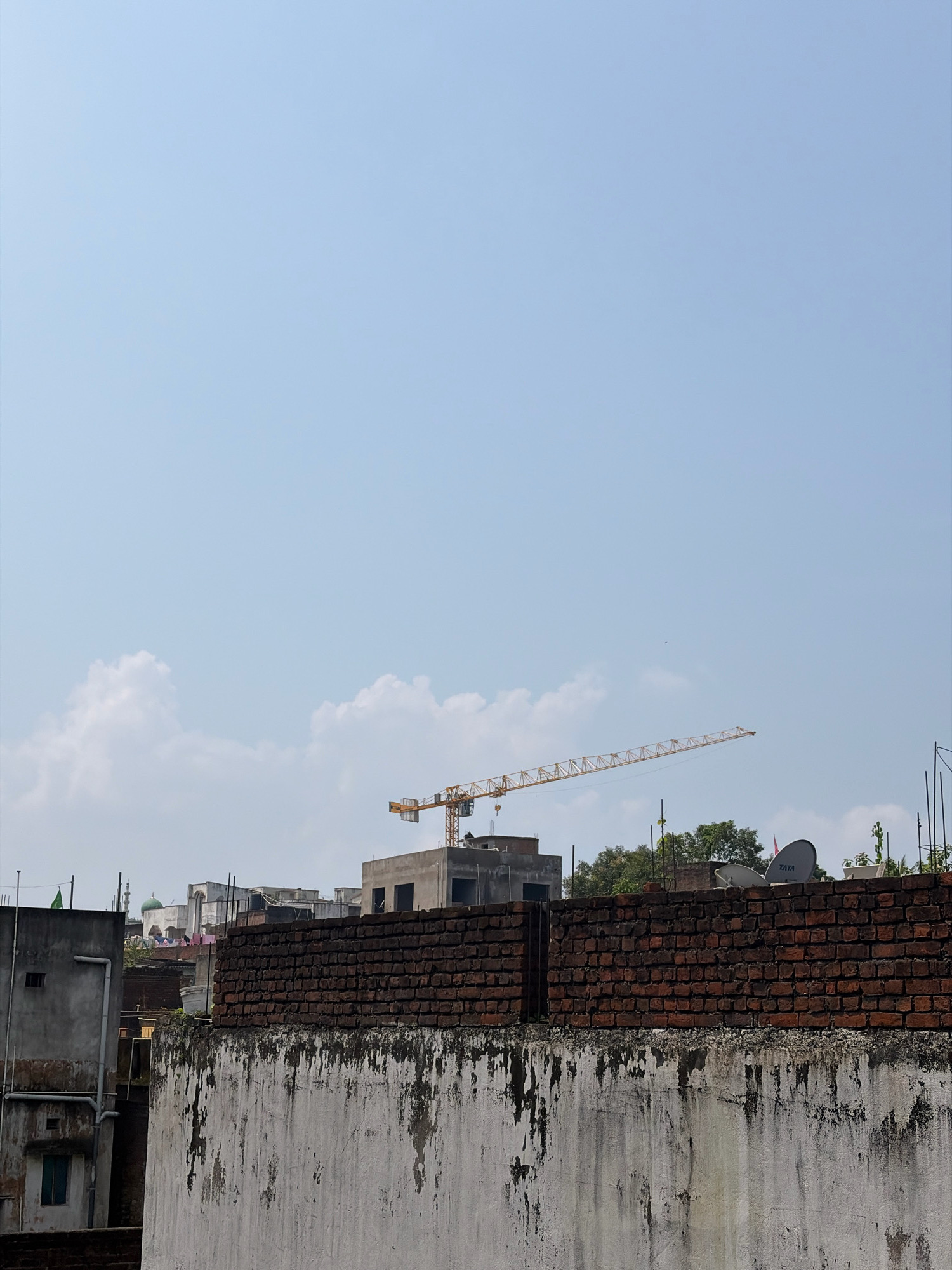 This is a street-level, medium-shot photograph taken in the city of Jamshedpur, India, under a clear, bright sky with scattered white clouds. The foreground is dominated by a weathered, white-plastered wall with significant water stains and patches of exposed brick, which is topped by a dark brick wall. Beyond the foreground wall, a complex urban landscape unfolds.
In the mid-ground, several buildings of varying heights and construction materials are visible. To the left, a gray concrete building with a simple window and a vertical pipe running down its facade is present. Adjacent to it, another building constructed with dark bricks is partly obscured. Further into the mid-ground, a mosque with a green dome and a minaret stands out against the sky, suggesting the presence of a religious site. Other buildings, some appearing to be under construction or in a state of disrepair, are scattered across this section. Clothes are seen hanging on a line in front of one of these buildings.
The background features a prominent yellow construction crane extending diagonally across the frame, indicative of ongoing development. Two satellite dishes are visible on the roof of a building in the right mid-ground, one bearing the "TATA" logo. Dense green trees are interspersed with the buildings, adding a touch of nature to the urban scene. The overall impression is one of a densely populated urban area with a mix of older and newer constructions, characteristic of many Indian cities like Jamshedpur. No people are visible in the frame.