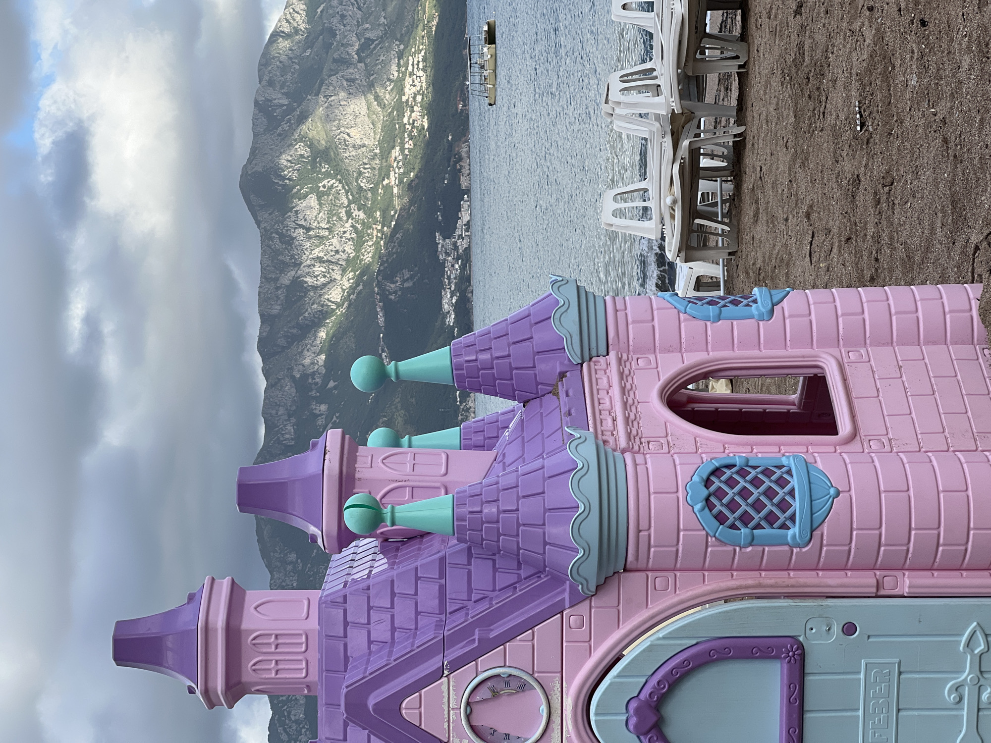 The image shows a child's pink and purple plastic castle playhouse on a sandy beach, with a backdrop of the sea, mountains, and a cloudy sky. White plastic chairs are stacked near the playhouse. The location is likely in Budva, Montenegro, considering the landscape and architecture visible.
