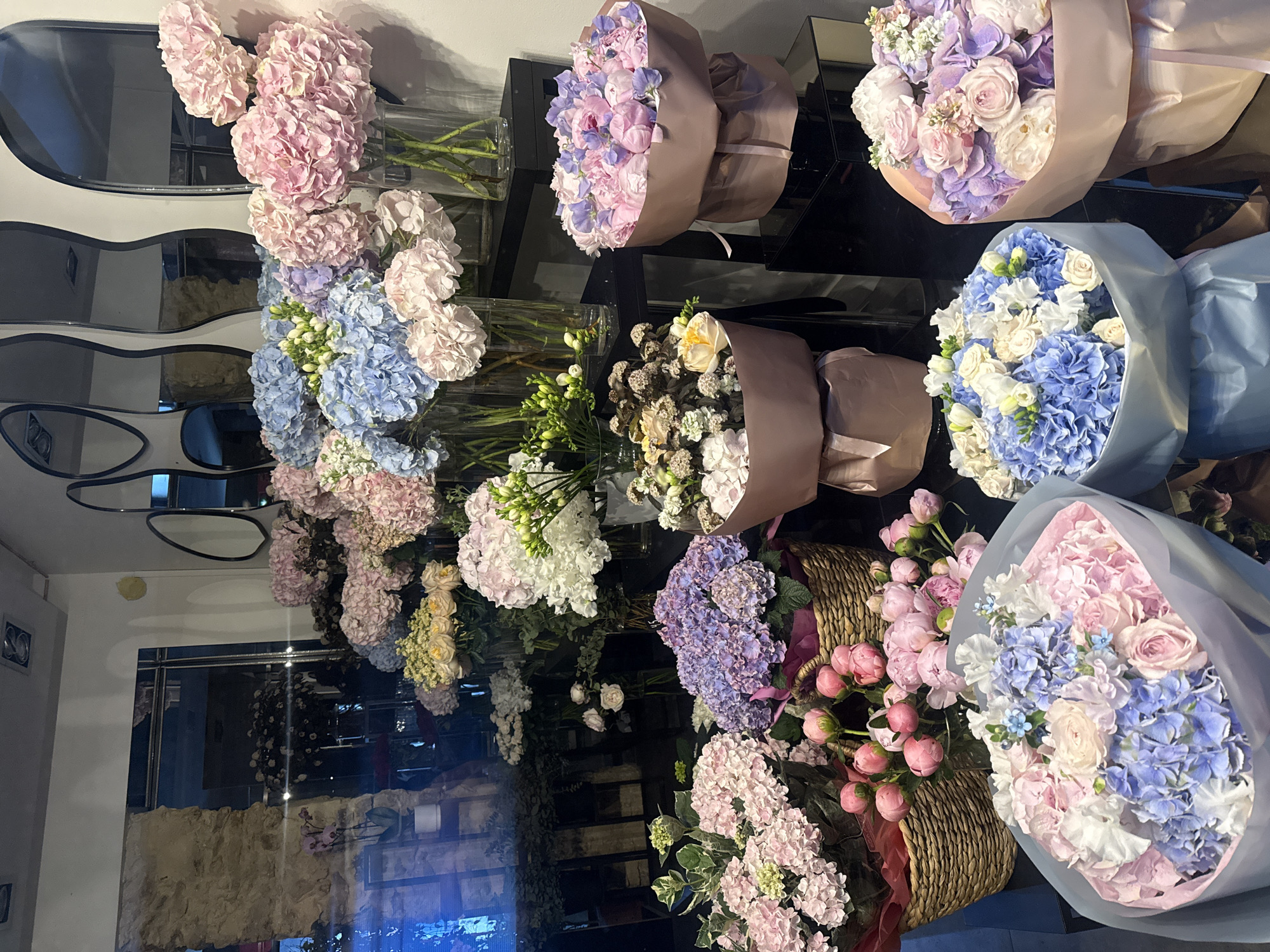The image depicts a flower shop interior. There are multiple bouquets and floral arrangements. Some arrangements are in glass vases, while others are wrapped in paper or placed in wicker baskets. The flowers are a variety of colors including pink, purple, blue, and white. The shop appears to have a reflective surface, possibly a black countertop, which mirrors the flowers. Based on the floral styles and arrangements, it is likely the shop is located in an urban area, potentially Paris, France, due to the sophisticated floral design often associated with Parisian florists. No people are visible in the frame.