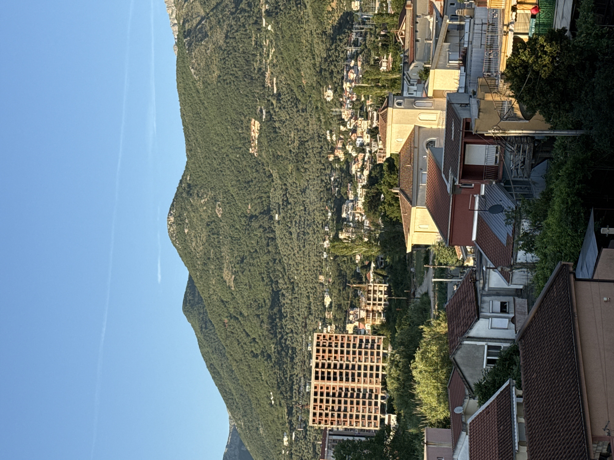 The image presents a scene from Bar, Montenegro, showing a hillside covered in dense green forest that slopes upwards toward a clear blue sky. Streaks resembling contrails are visible in the sky. Nestled amongst the trees on the hillside are numerous residential buildings, appearing as a scattered settlement. Closer to the foreground, there is a partially constructed, rectangular building with a grid-like frame. Adjacent to this structure are various rooftops, suggesting the presence of residential houses with brown and gray roofs. The overall scene depicts a natural landscape integrated with urban development.