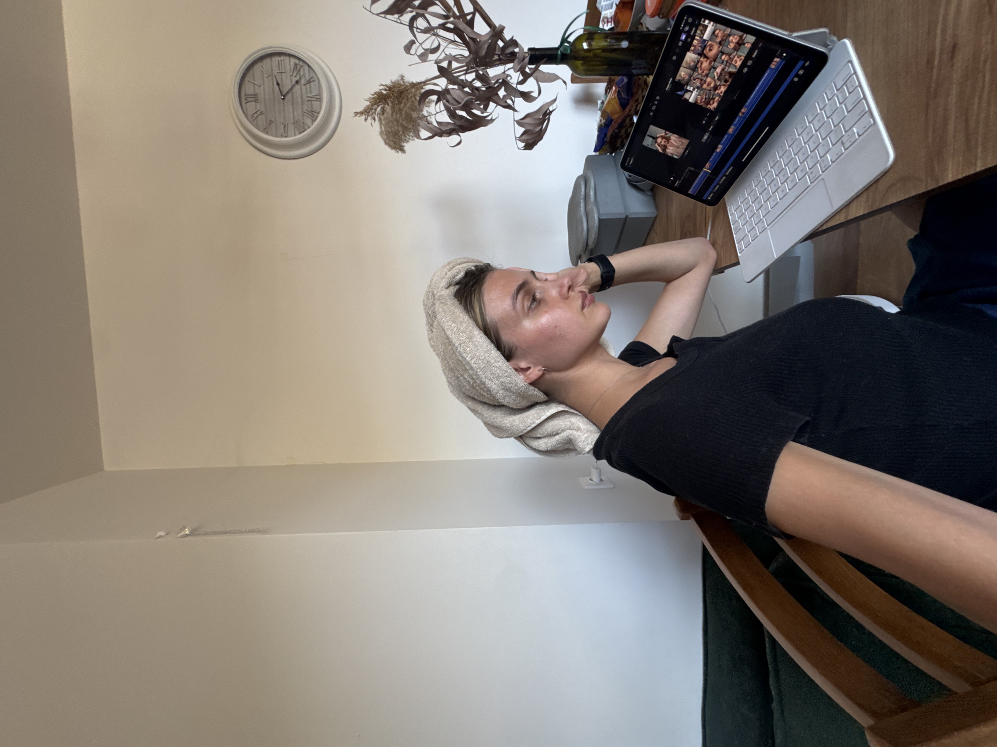 A young woman is depicted indoors, reclining or lying down with a towel wrapped around her head, indicating a recent shower or skincare routine. She wears a black, ribbed top and appears to be looking towards an iPad positioned on a wooden table to her right. Her right arm is bent with her hand resting near her head, suggesting a relaxed or contemplative posture.
On the wooden table, an iPad with an attached light-colored keyboard is open, displaying what appears to be video editing software or a gallery of video clips, characterized by multiple small preview thumbnails and a timeline. Behind the iPad, a dark green bottle holds dried plants or foliage. A light grey cylindrical object, possibly a speaker or humidifier, is also on the table.
On the plain, light-colored wall behind her, a large, round white clock with Roman numerals marks the time as approximately 1:55. The overall setting suggests a casual home environment. Part of a wooden chair and a dark cushion are visible in the lower right corner, blending into the scene. The lighting appears natural and soft. The woman's expression is calm and thoughtful.