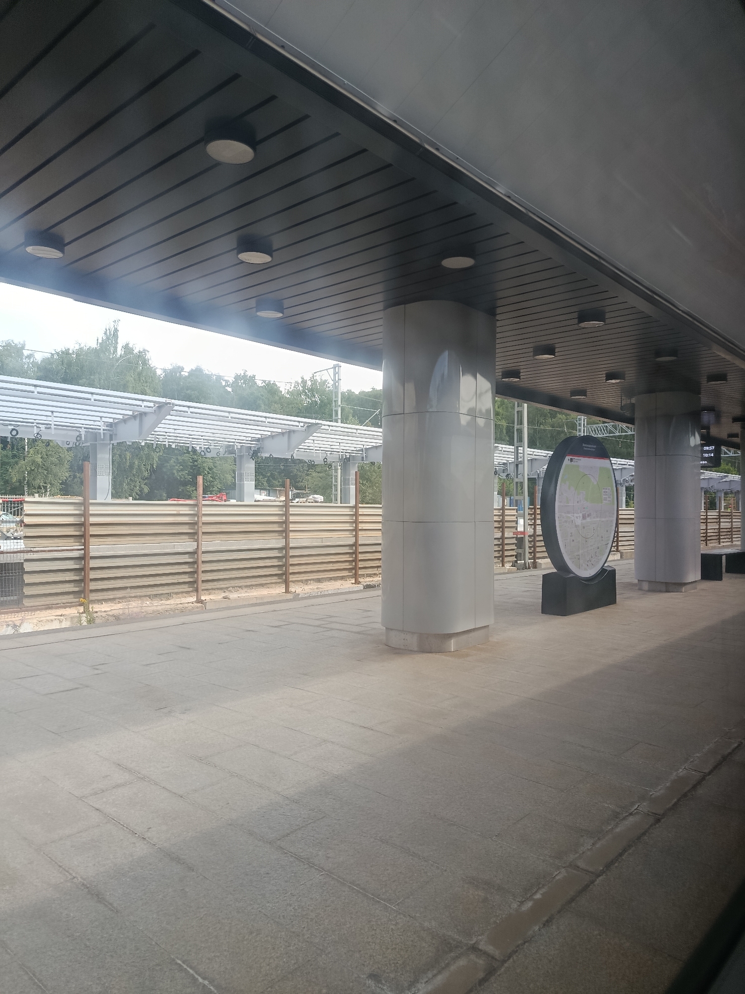 The image shows a covered outdoor public space, possibly a train station platform in Balashikha, Russia. The ceiling is a dark gray, ribbed structure with multiple circular light fixtures. Several large, light gray cylindrical pillars support the roof. To the right, a large, circular map display is situated on a dark rectangular base. The map itself is a detailed diagram with text in Russian. Beyond the pillars, a light-colored tiled floor stretches into the distance. In the background, there is a long, light-colored horizontal fence made of layered panels, supported by vertical brown posts. Above the fence, a metal structure with multiple horizontal beams is visible, possibly part of a larger railway infrastructure. Trees and foliage are present in the far background, suggesting an outdoor environment.