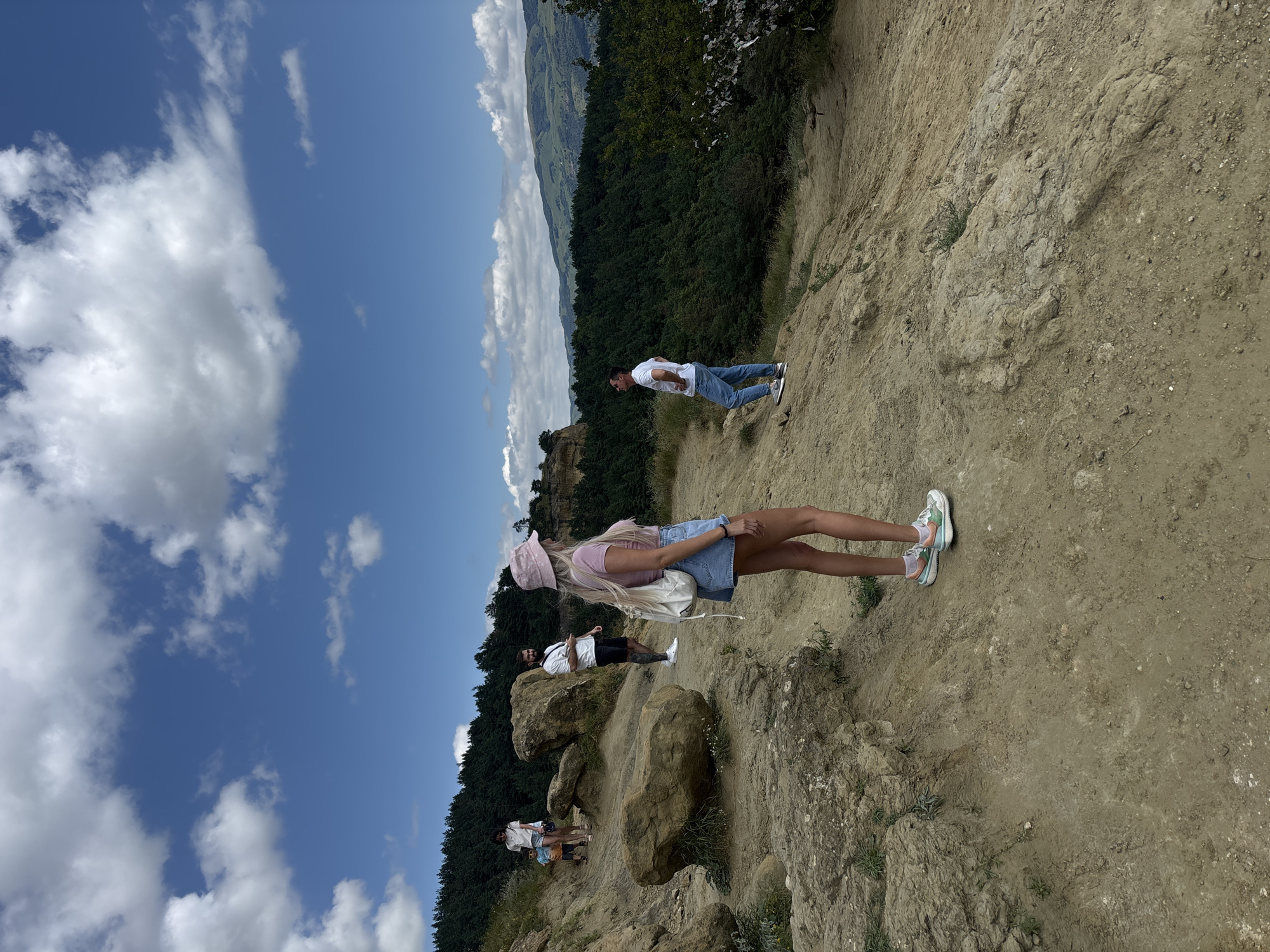 The image shows a group of people on a dirt path on a hillside overlooking a forest. The sky is blue with some white clouds. In the foreground, a woman with long blonde hair wearing a pink bucket hat, a pink t-shirt, and denim shorts is facing away from the camera, looking out at the view. She has a white backpack on. Behind her and to her left, a man with a beard, wearing a white t-shirt and black shorts, is looking at his phone. Further back and to the left, a woman in a white t-shirt and shorts is walking with a young boy. To the right of the frame, a man in a white t-shirt and blue jeans is looking up at the sky. The hillside is covered in dry grass and rocks, with some larger boulders. The background is dominated by a dense green forest that extends to the horizon, where rolling hills can be seen under a cloudy sky. There is no visible indication of the city of Mirny, Russia in this image.