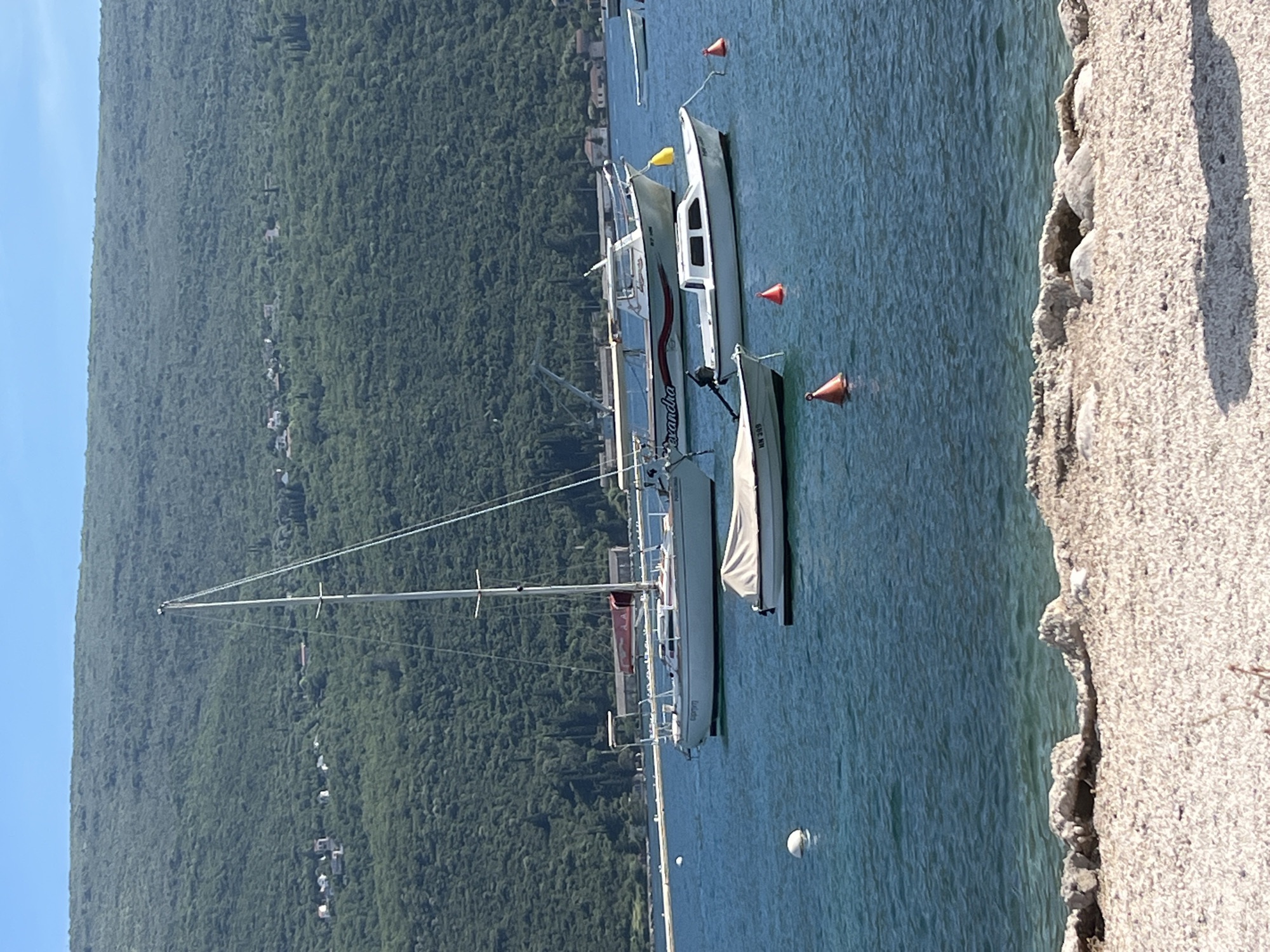 The image displays a serene coastal scene in Đenovići, Montenegro, on a bright, sunny day. In the foreground, a light-colored, textured stone quay or dock runs vertically along the right side of the frame, indicating a shoreline or harbor edge.
The midground is filled with calm, blue-green water, where several boats are either moored or anchored. A prominent white sailboat with a tall mast and a small red feature on its hull is visible towards the left of the water. To its right, closer to the quay, two smaller boats are present. One is a white motorboat with red and black stripe details, clearly displaying the name "Alexandra" on its side. Adjacent to this, a smaller, light-colored dinghy or tender is seen, with the registration number "HN 369" marked on its bow. Several orange and white spherical buoys are scattered across the water.
In the background, a large, verdant mountain or hill rises dramatically, covered in dense green foliage. A line of small, light-roofed houses is visible nestled along the mountain's slope, indicating a small settlement. The sky above is a clear, bright blue, suggesting fair weather and abundant sunshine. The scene captures a tranquil moment of boats resting on the water against a picturesque mountainous backdrop.
