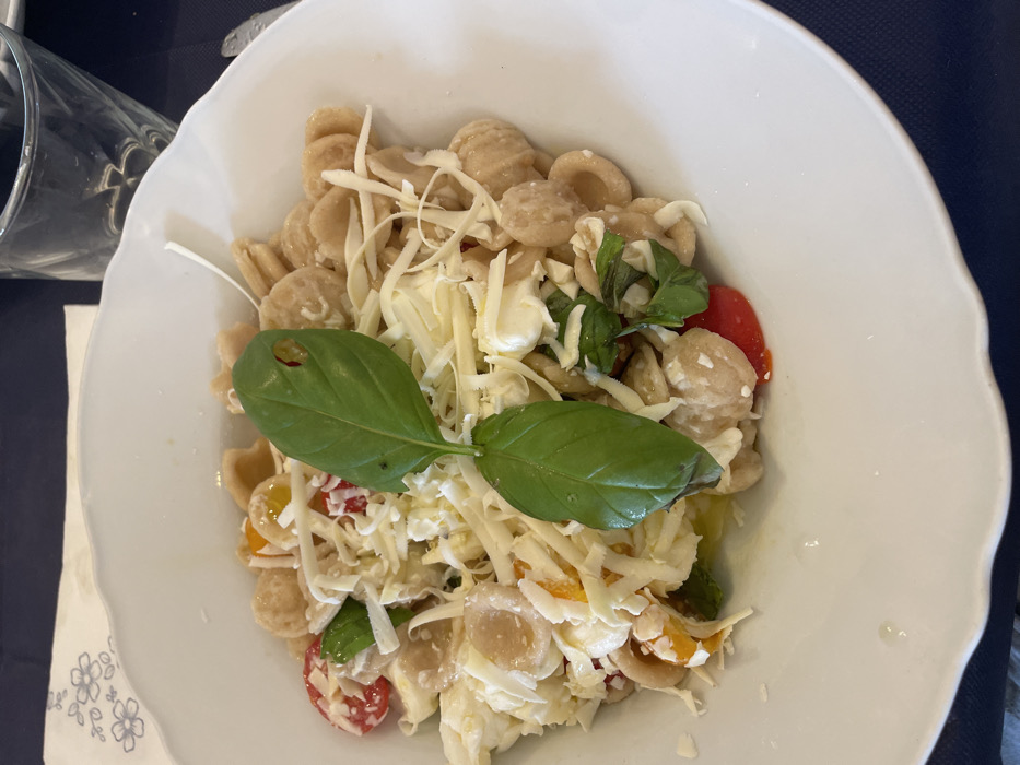 A white scallop-edged bowl, located in Ceglie Messapica, Italy, contains a pasta dish. The dish is composed of light brown orecchiette pasta, topped with shredded white cheese, small cubes of fresh mozzarella, halved red and yellow cherry tomatoes, and fresh green basil leaves. One prominent basil leaf in the center displays a small, irregular perforation. A subtle sheen consistent with olive oil is visible at the bottom of the bowl. The bowl rests on a dark blue tablecloth. A white paper napkin with a light blue floral pattern is partially visible to the upper left of the bowl, and a clear glass of water is partially visible to the upper right. A silver utensil handle is barely visible on the far right edge. No individuals or direct actions are depicted.