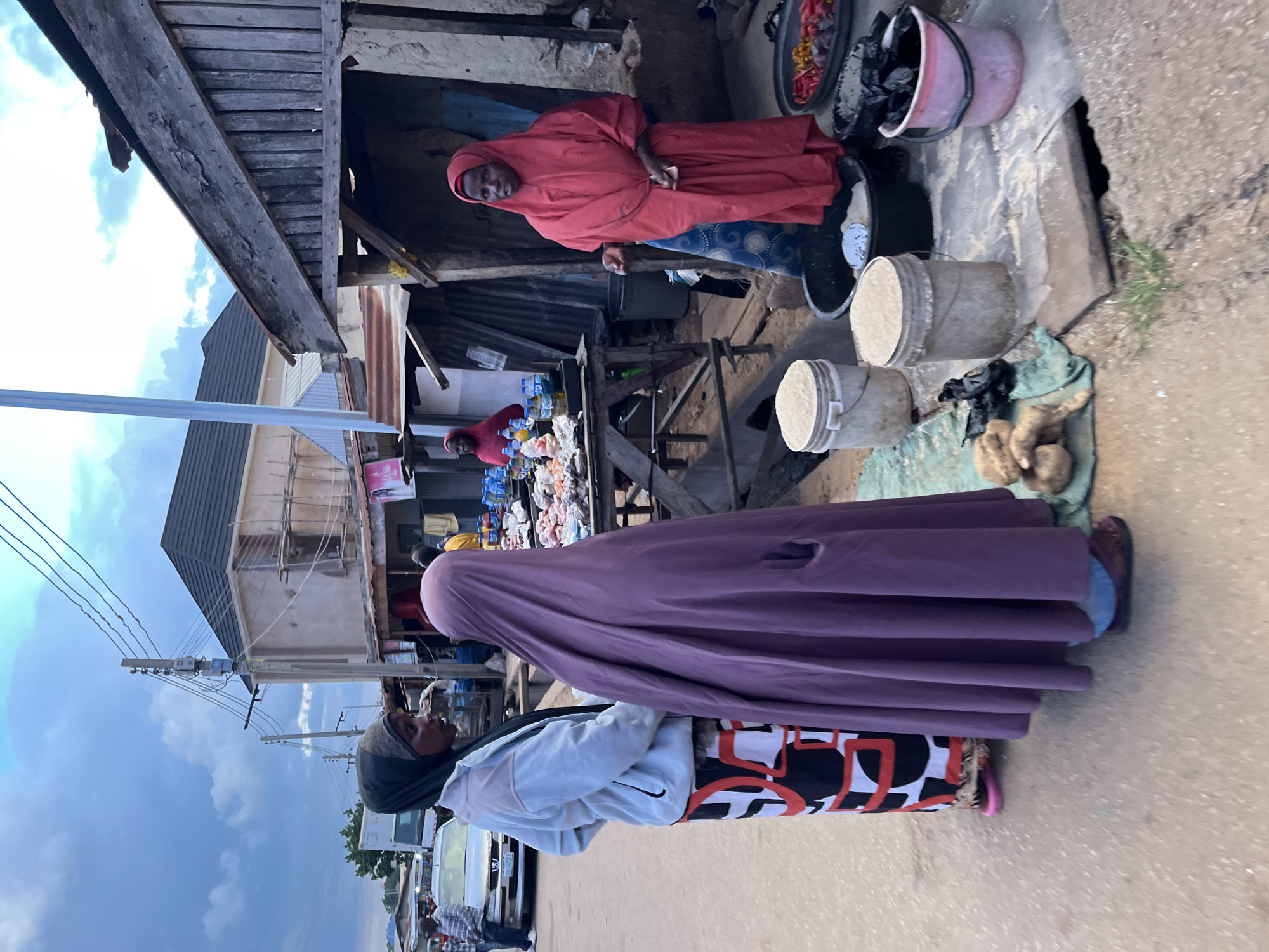 This street scene in Jos, Nigeria, captures a bustling outdoor marketplace or street vendors. The image, rotated 90 degrees, shows a cloudy sky overhead, suggesting either an overcast day or late afternoon.
Three women are prominent subjects. In the foreground, a woman wears a dark head covering, a light-colored long-sleeved top with a black logo (resembling a Nike swoosh) on the chest, and a patterned skirt featuring black, white, and red geometric shapes. Over this, she drapes a long, plain purple garment. She looks towards the left side of the frame.
In the midground, another woman stands behind a wooden stall, dressed in a red head covering and matching long-sleeved top, blue patterned pants, and sneakers. She looks directly at the viewer with a neutral expression. Her stall displays numerous clear plastic bottles, possibly containing beverages, and other items. Further back and to the left, a third woman, similarly dressed in a red head covering and garment, tends to another stall with goods.
The ground in the foreground shows various items for sale or storage, including several buckets of different colors (white, grey, pink, black), some containing what appears to be granular material. Root vegetables, possibly yams, rest on a piece of fabric. The stalls themselves are constructed from rough wooden planks and corrugated metal sheets, indicating a simple, makeshift market setup.
In the background, other buildings are visible, along with utility poles and overhead power lines, contributing to the urban or semi-urban street atmosphere. The overall scene depicts daily commerce and street life.
