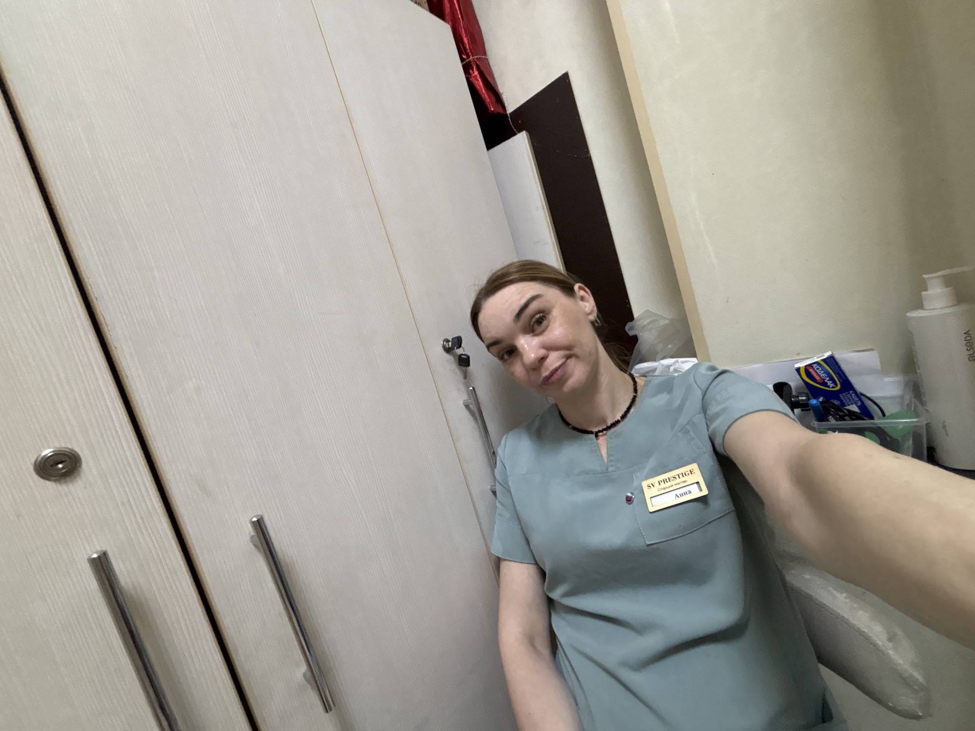 A female individual, identified by a name tag as Anna, a Senior Master at "SV PRESTIGE," is depicted indoors within a professional establishment in an unknown city in Russia. Anna, with light brown hair, wears a light teal short-sleeved scrub top and a dark beaded choker. Her name tag is yellow with black text and a small red and white emblem. She is captured in a selfie, holding the camera with her right arm, which is partially visible. Her expression is a subtle smile.
The immediate background to Anna's left consists of two light-colored wooden cabinet doors with vertical grain, equipped with silver-toned tubular handles and a single silver keyhole on the left door. Behind Anna to her right, a white wall or partition transitions into a darker brown panel, followed by a lighter cream-colored wall. Various items are visible on a surface to her right: a white pump bottle, a clear plastic container holding a blue and yellow box labeled "КОДЕЛАК" (KODELAK - a Russian cough medicine), and other miscellaneous supplies. A crumpled red foil or plastic object is positioned in the upper right background. Part of a light-colored, textured armrest, possibly from a chair or couch, is visible in the lower right corner of the frame. The lighting appears to be artificial.