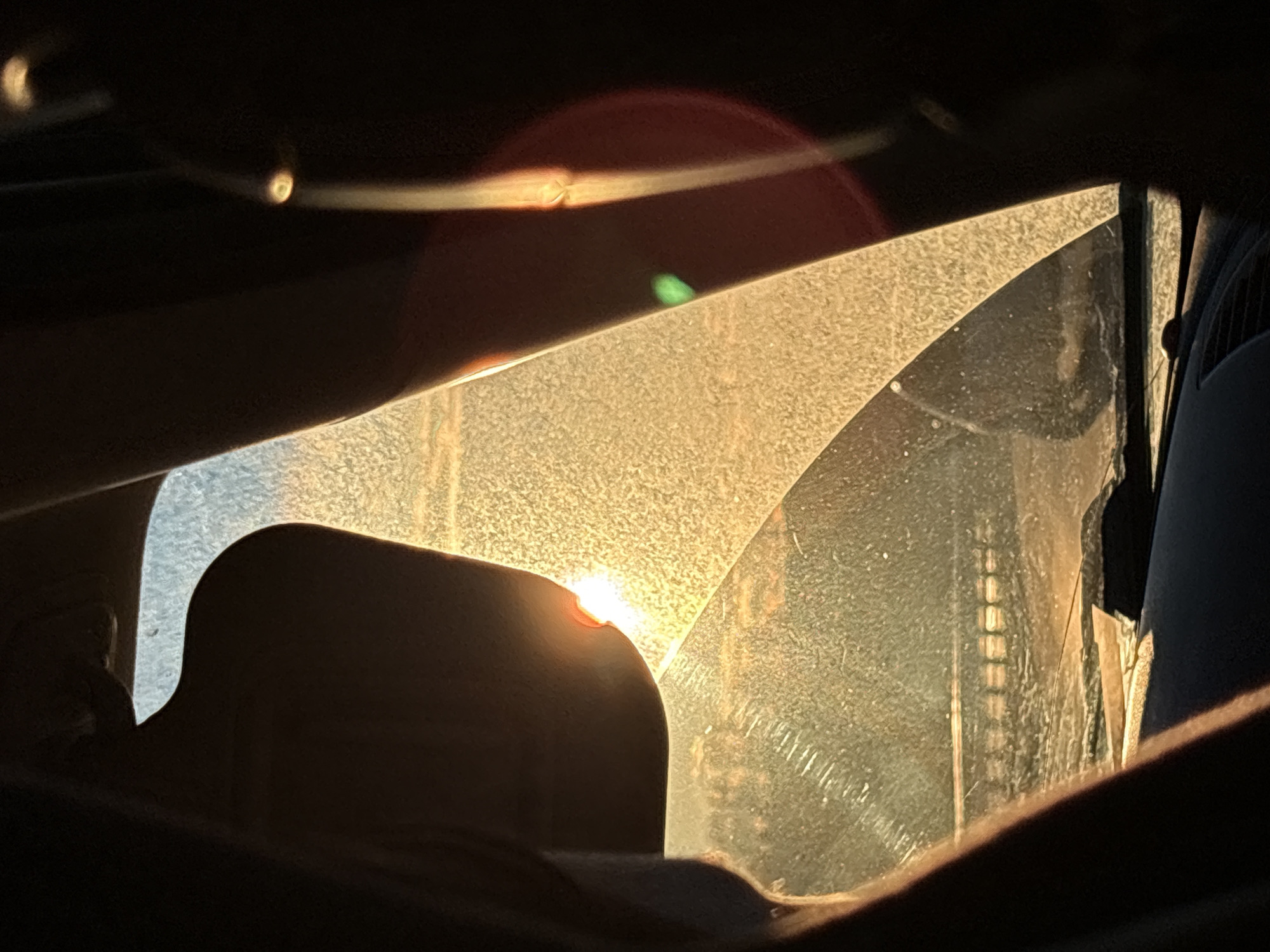 The image captures the interior of a vehicle, likely a car, heavily illuminated by strong, direct sunlight. The scene is characterized by high contrast, with significant portions of the car's interior appearing in deep silhouette, primarily the dashboard or door panel in the foreground and around the illuminated window.
The main subject is a section of a car window, possibly the windshield or a side window, which is brightly lit by the sun. This glass surface appears to be quite dirty or dusty, causing the sunlight to scatter and create a textured, hazy effect across its surface. A small, intense point of light, presumably the sun itself, is visible peaking over a silhouetted edge within the frame, casting a warm glow.
Prominent lens flares are visible, including a large reddish halo and a smaller, distinct green elliptical flare, suggesting the sun is directly hitting the camera lens. Through the illuminated glass, some faint, vertical structural elements are discernible in the background, possibly reflections or distant objects outside the vehicle like a fence or building. The strong, low-angle sunlight indicates either early morning or late afternoon. No specific activity or people are depicted. There is no visible text in the image. The location context provided is Safonovo, Russia.