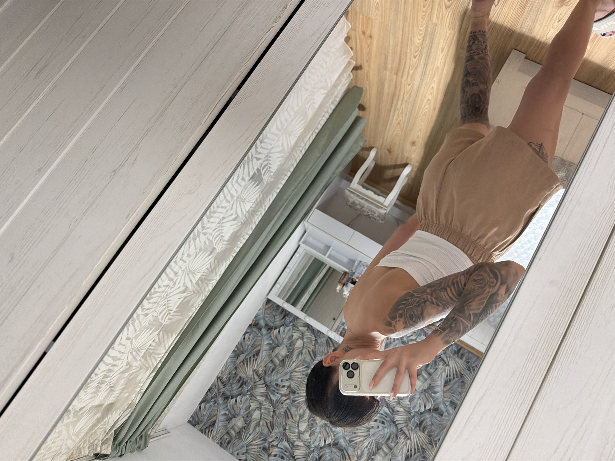 A woman is taking a mirror selfie in what appears to be a bedroom. She is wearing a white tank top and beige shorts. She has short, dark hair and visible tattoos on her arms and legs. The room features a vanity with a mirror, a small stool, and patterned wallpaper. The floor is wooden, and the overall lighting is soft. The image is not taken in Plovdiv, Bulgaria.