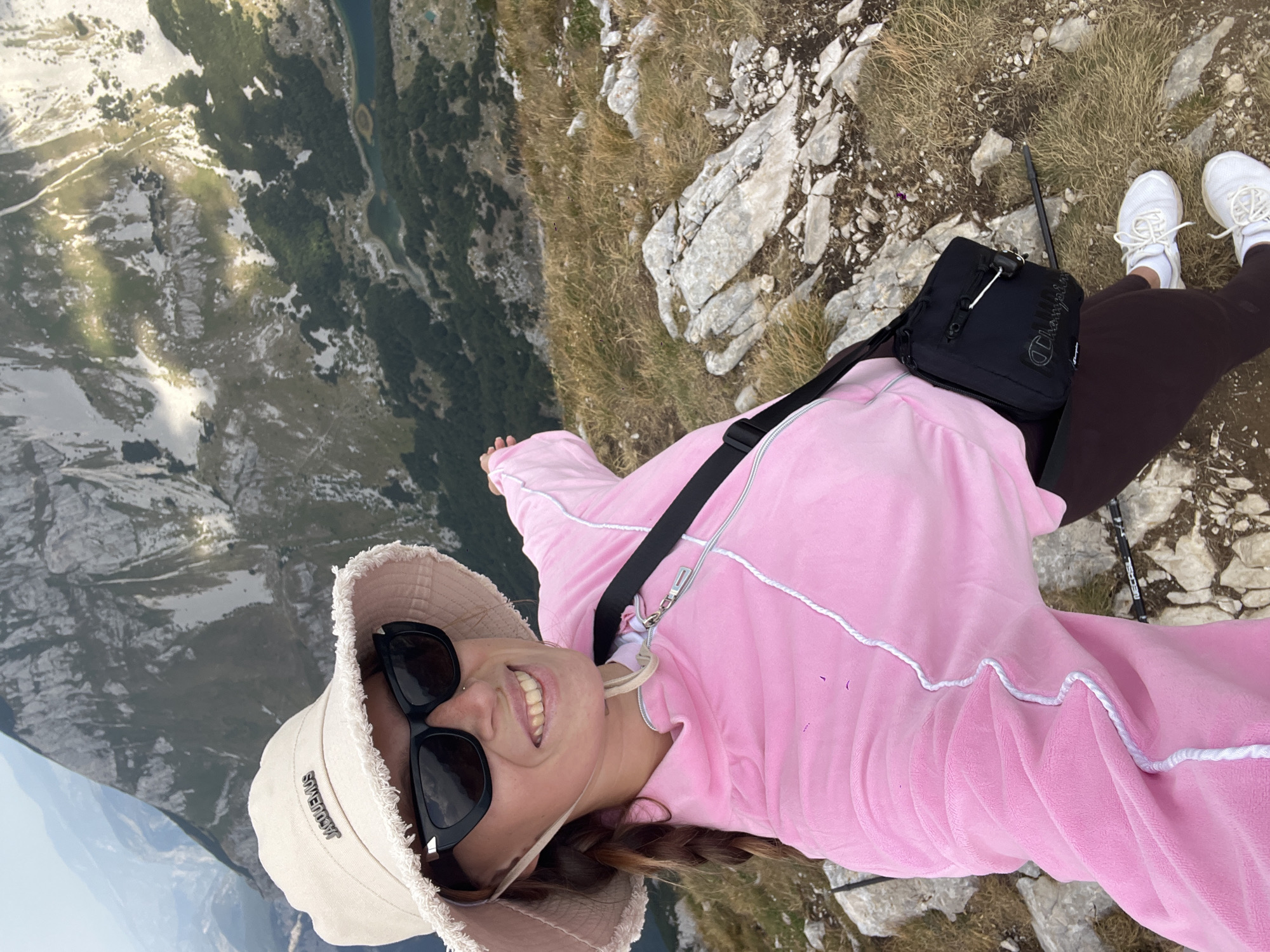 A young woman with braided brown hair, wearing a pink velour tracksuit with white trim, a beige bucket hat with "JACQUEMUS" branding, black sunglasses, and a black crossbody bag, stands on a rocky, grassy mountain slope. She is smiling and pointing towards the vast mountainous landscape of Durmitor National Park near Боричје, Montenegro. The background reveals a breathtaking vista of rugged peaks, dense green forests, and a serene blue lake. The lighting suggests it is daytime, with some sunlight filtering through the clouds, casting shadows on the mountainside. The woman's attire indicates she is dressed for outdoor activity.
