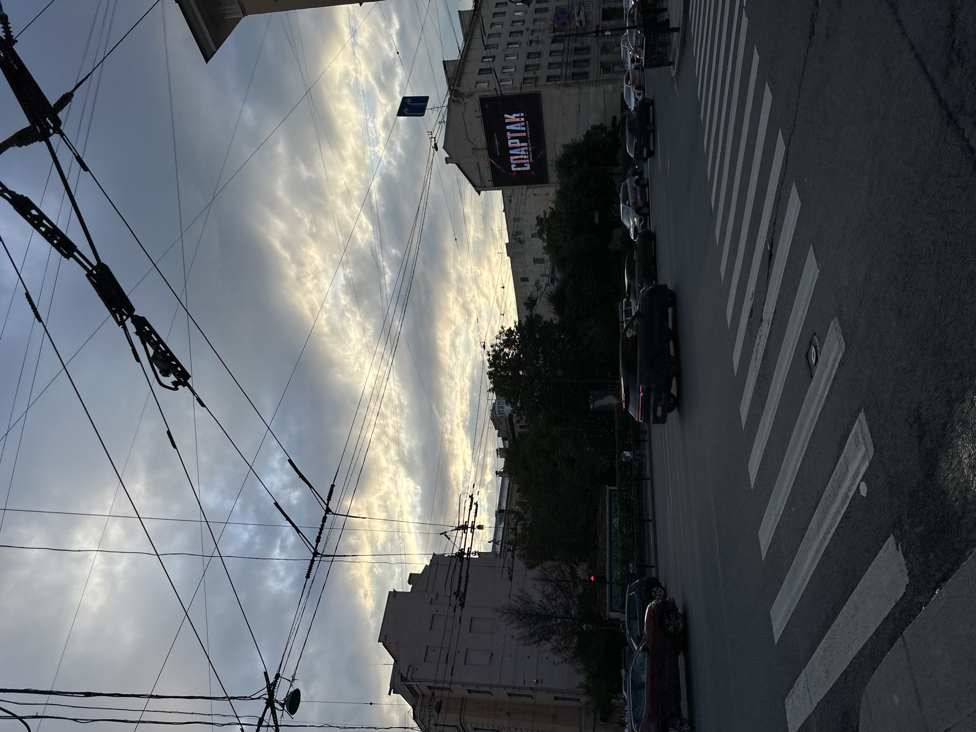 This image depicts a street scene in Saint Petersburg, Russia, likely during twilight given the lighting conditions and the cloudy sky. The foreground features a crosswalk with several vehicles, including SUVs and sedans, driving on the asphalt road. Overhead, a complex network of trolleybus wires crisscrosses the sky, a characteristic feature of the city's public transportation system.
In the mid-ground, a large billboard is prominently displayed on the side of a multi-story building, advertising "СПАРТАК" (Spartak), with what appears to be event details or a title below it. The building itself is typical of Saint Petersburg's architecture, with multiple windows and a somewhat aged facade. Adjacent buildings, trees, and streetlights are also visible. The overall atmosphere is urban and functional, with a sense of everyday city life unfolding under a dramatic sky. No specific individuals are clearly identifiable or engaged in any particular action.