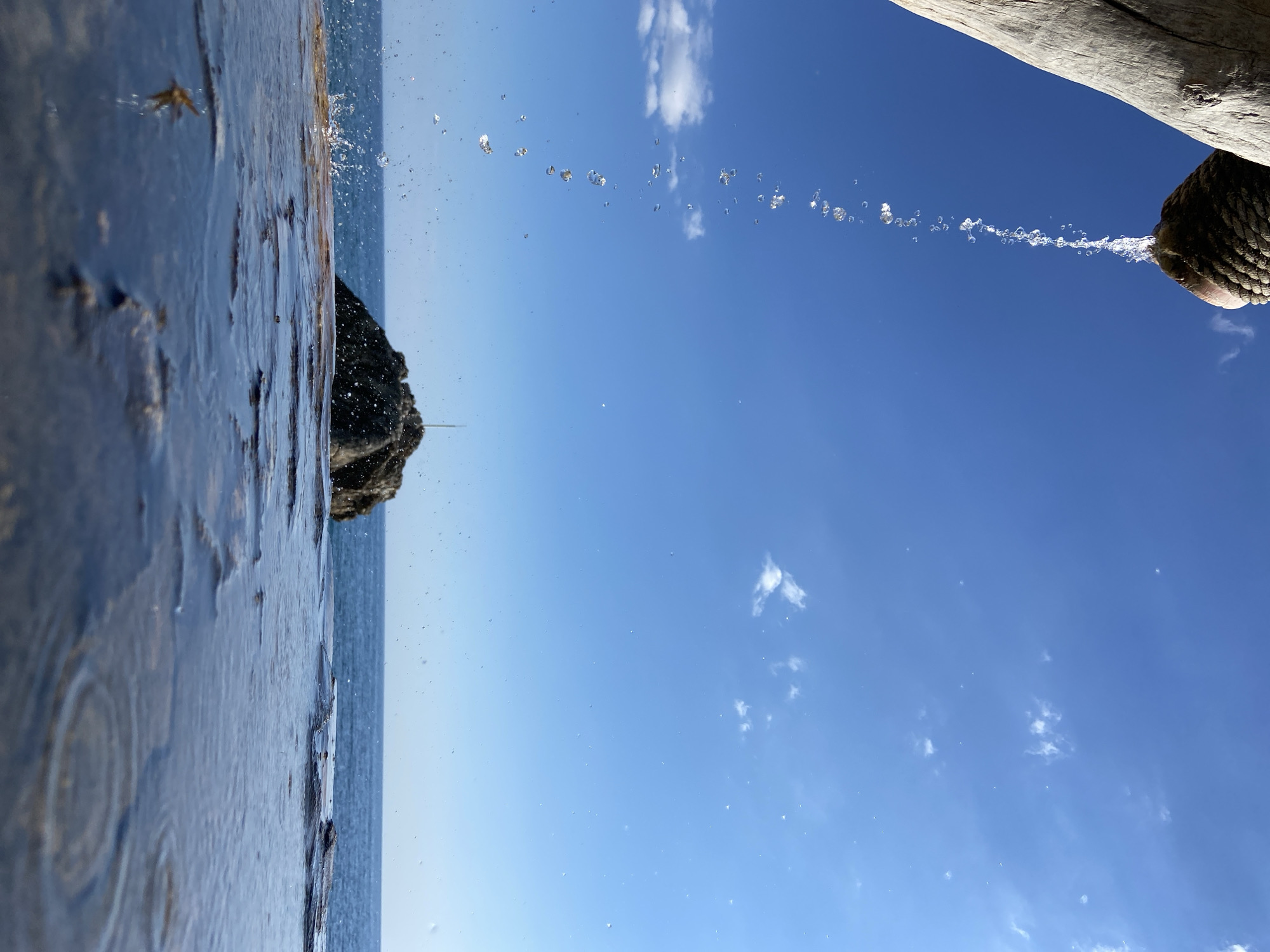 Captured at Prijevor, Montenegro, the image portrays a water fountain set against a backdrop of a clear blue sky with scattered clouds. Water gushes from a rustic wooden spout, its flow frozen mid-air into a series of distinct droplets. In the background, a dark, rocky outcrop rises from the sea, partially obscured by the water. The foreground is dominated by a reflective wet surface, possibly a shallow pool or pavement, mirroring the sky and adding depth to the scene.
