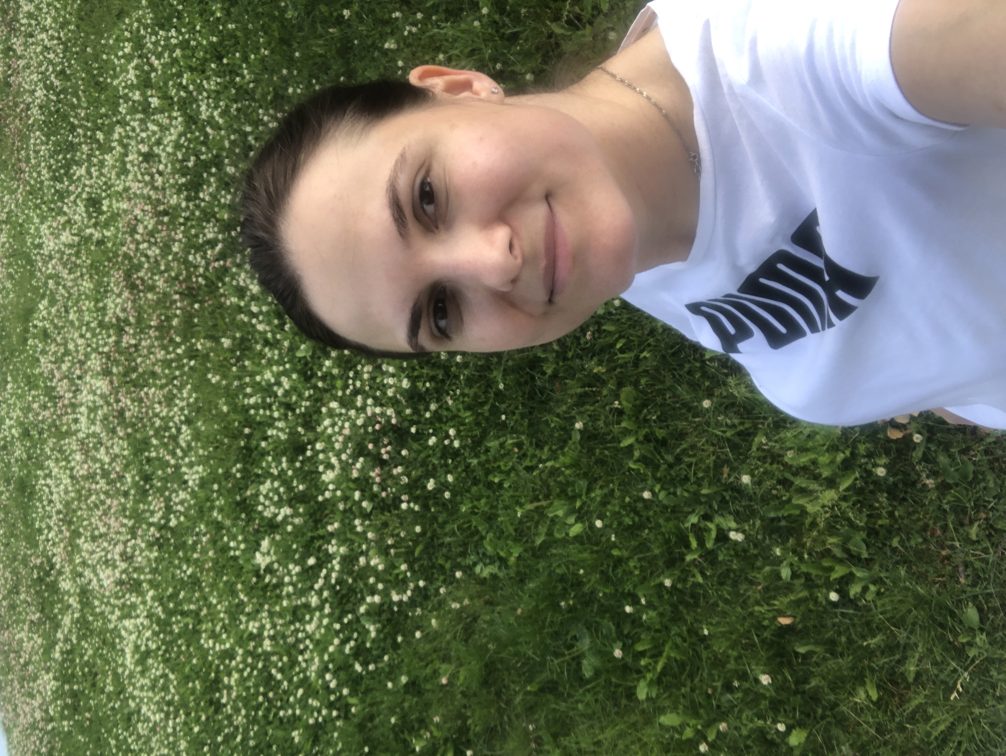 A woman is lying down on a patch of grass dotted with small white flowers. She is taking a selfie and smiling at the camera. Her hair is dark and pulled back from her face. She is wearing a white t-shirt with a black Puma logo on it, and a thin silver necklace. The grass is green and lush, indicating it is likely springtime or summer. The image appears to be taken outdoors in daylight, with no specific time of day or weather conditions discernable. The setting is a natural outdoor environment, possibly a park or a backyard, in Moscow, Russia. The woman's expression is cheerful and relaxed.