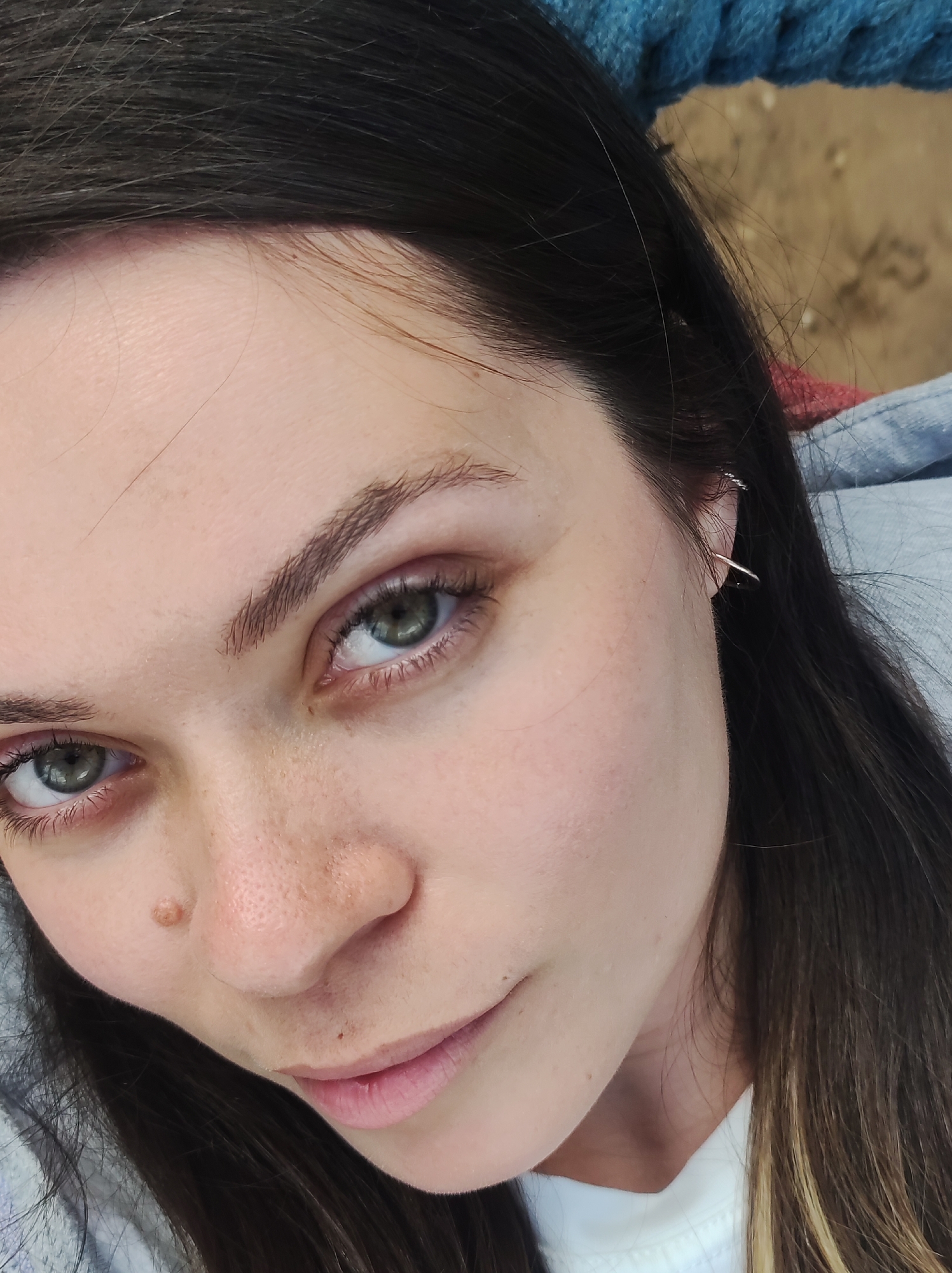 Close-up selfie of a Caucasian woman with dark hair and green eyes. She has a mole on her left cheek near her nose. She is wearing a silver earring in her left ear. Her lips are slightly parted in a subtle smile. She is wearing a white shirt and a blue denim jacket, with a hint of blue cable knit behind her head. The background appears to be sand, suggesting an outdoor setting. The image was not taken in the city of Chaikovsky, Russia, according to the prompt.
