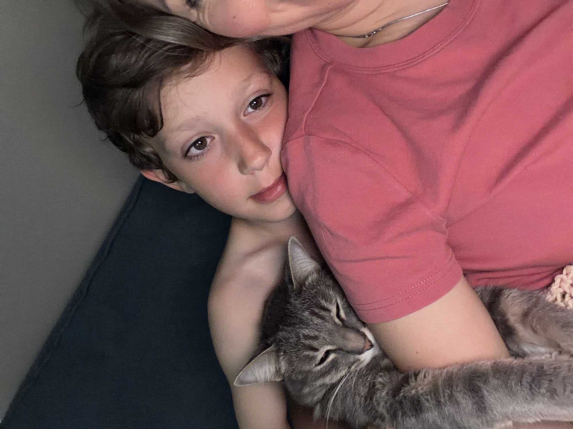 A photograph shows a close-up view of a child and an adult, along with a cat. The child is likely lying down, with their face close to the adult's. The cat is being held in the adult's arms. The adult is wearing a red shirt. The location is unknown, there is no definitive evidence of Chaikovsky, Russia.