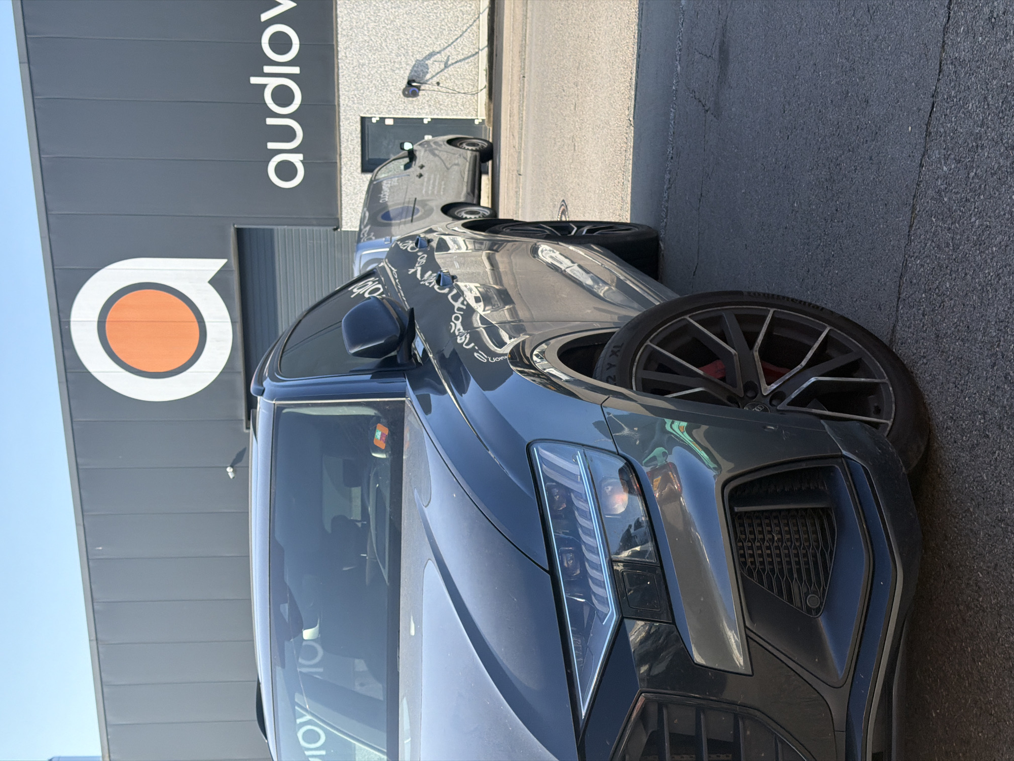 This is a low-angle, medium shot of a dark gray SUV parked in front of a modern building. The building's facade is dark gray with vertical panels and a large white logo featuring an orange circle within a speech bubble shape. To the right of the logo, white text reads "audiov." A light gray commercial van with a similar logo and text is parked behind the SUV. A charging station with a cable is attached to the side of the building. The SUV's front end is in the foreground, with its headlights and grille visible. The wheels of the SUV are black with silver accents and red brake calipers. The ground is paved with asphalt. The sky is clear and blue. The image appears to be taken in Leudelange, Luxembourg, based on the visible signage.