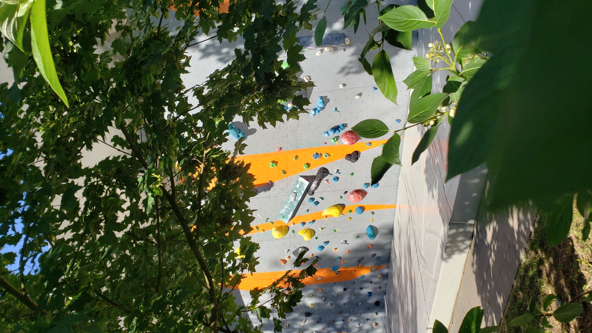 A bright, outdoor climbing wall is visible, partially obscured by lush green foliage in the foreground and dappled sunlight filtering through the leaves. The wall itself is grey with large, striking orange diagonal stripes. Numerous colorful climbing holds in shades of blue, yellow, red, black, and grey are scattered across the wall, indicating various climbing routes. Some of the holds are unusually shaped, including rounded, bulbous ones, and flatter, rectangular ones. There is a large, padded mat at the base of the wall, a common safety feature for climbing areas. The ground in front of the mat appears to be a mix of grass and paved or tiled surfaces, with shadows cast from the surrounding trees. The image was taken in the city of Prešov, Slovakia, specifically at the Lúčka Bouldering Gym, located at Ulica Lúčna 11. The scene suggests a sunny day, suitable for outdoor recreational activities.