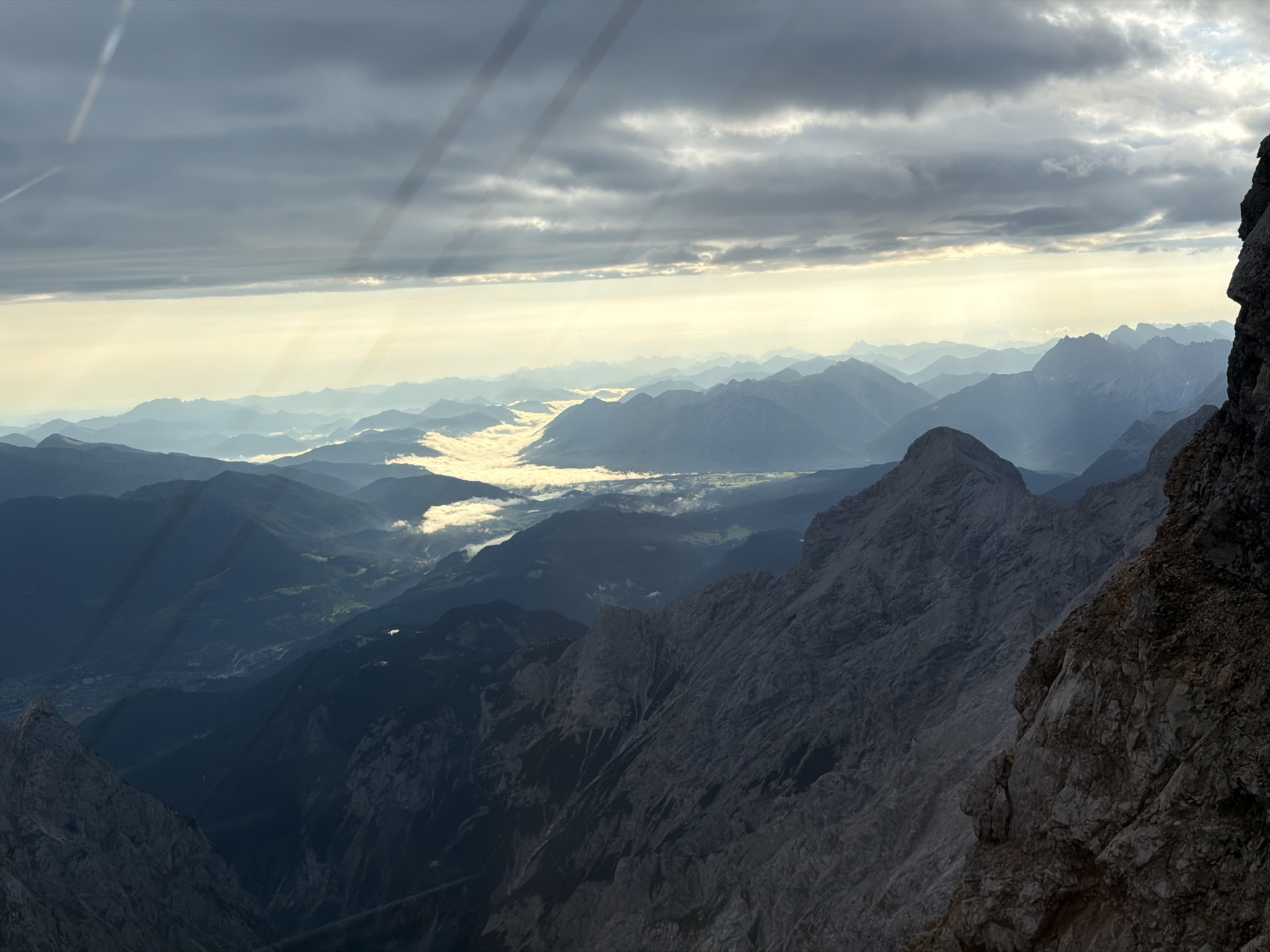 This is a panoramic, high-angle photograph capturing a mountain range at dusk. The foreground is dominated by a sheer, rocky cliff face on the right side, casting a shadow over the adjacent mountain slopes. In the mid-ground, a series of dark, imposing mountain peaks recede into the distance, their sharp ridges silhouetted against the sky. A valley floor, filled with a layer of soft, white clouds, is visible between the mountains, creating a striking contrast between solid earth and ethereal vapor. The background showcases a vast expanse of layered mountain ranges, fading into a hazy blue due to atmospheric perspective. The sky above is dramatic, with dark, heavy clouds interspersed with shafts of golden sunlight breaking through, illuminating the landscape below with a warm, ethereal glow. The overall scene suggests a remote, high-altitude environment, likely part of the Bavarian Alps near Grainau, Germany, given the characteristic alpine terrain and the quality of light often seen in this region at twilight. There are no people or man-made structures directly visible in this natural landscape.