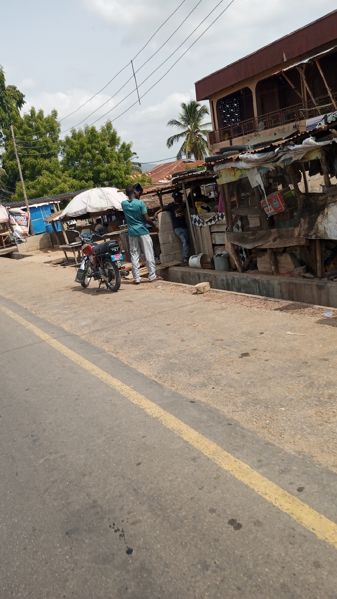The photo, taken during daylight in Zango-ndaji, Nigeria, shows a street scene. In the foreground is a paved road with a yellow dividing line. A person wearing a turquoise shirt and light-colored pants stands on the sidewalk next to a black motorcycle. This person appears to be looking or gesturing towards a ramshackle storefront adjacent to them. Another individual wearing a black shirt and dark pants leans against the storefront. The store appears to be made of wood and corrugated metal. Above the storefront is a two-story building with a brown exterior and balconies. In the background, there are green trees and a cloudy sky, with overhead power lines visible. Other less-defined street stores are also in the background.