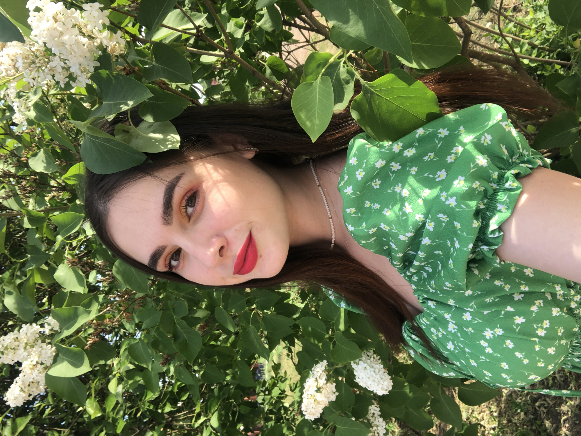 A young woman with dark brown hair is photographed amidst white lilac flowers and green leaves. She wears a short-sleeved green dress with a white floral pattern. Her makeup includes red lipstick and what seems to be a rose gold eye shadow. The leaves appear to be directly above her head and near her face, suggesting that the photo was taken at the level of a lilac bush. The sunlight suggests it is daytime, possibly outdoors. There is no indication this photograph was taken specifically in Noviy, Russia.