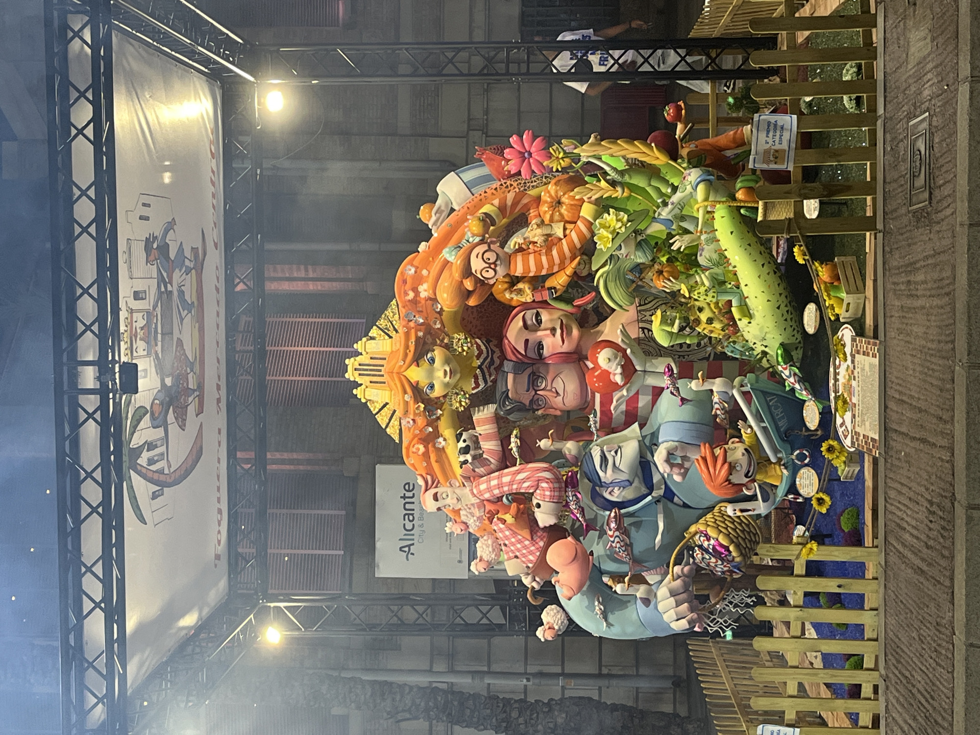 The image depicts a large, colorful, and intricately detailed monument, likely a Falla, displayed outdoors in Alicante, Spain. The monument is constructed from various materials and features numerous stylized figures, including humans, animals, and fantastical creatures, all arranged in a chaotic yet cohesive composition.
The monument is situated within a fenced-off area, marked by a light-colored wooden fence. Behind the monument, a building with shuttered windows is visible. Above the monument, a large banner hangs, displaying cartoonish figures and text that appears to be in Spanish. A sign reading "Alicante City & Beach" is also visible. The lighting suggests the scene is illuminated by artificial lights, likely for an evening event. A few people are visible in the background, one of whom is wearing a white t-shirt with text on it. The ground in front of the monument is paved. The overall impression is of a festive and artistic display, characteristic of local celebrations in Alicante.