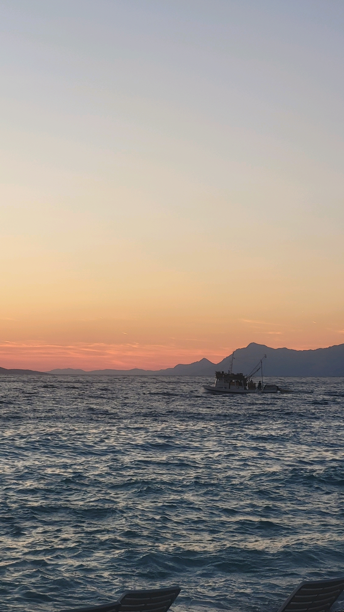 This image shows a body of water, likely the Adriatic Sea, with a sunset or sunrise in the background. A fishing boat is visible in the middle distance, and mountains can be seen on the horizon. In the foreground, the tops of two lounge chairs are visible, suggesting a beach or resort setting. The sky transitions from a pale blue at the top to warm orange and pink hues near the horizon. The water is choppy, with whitecaps indicating a moderate breeze. The overall impression is a serene coastal scene. The location is identified as Općina Tučepi, Croatia, a coastal municipality in the Split-Dalmatia County.