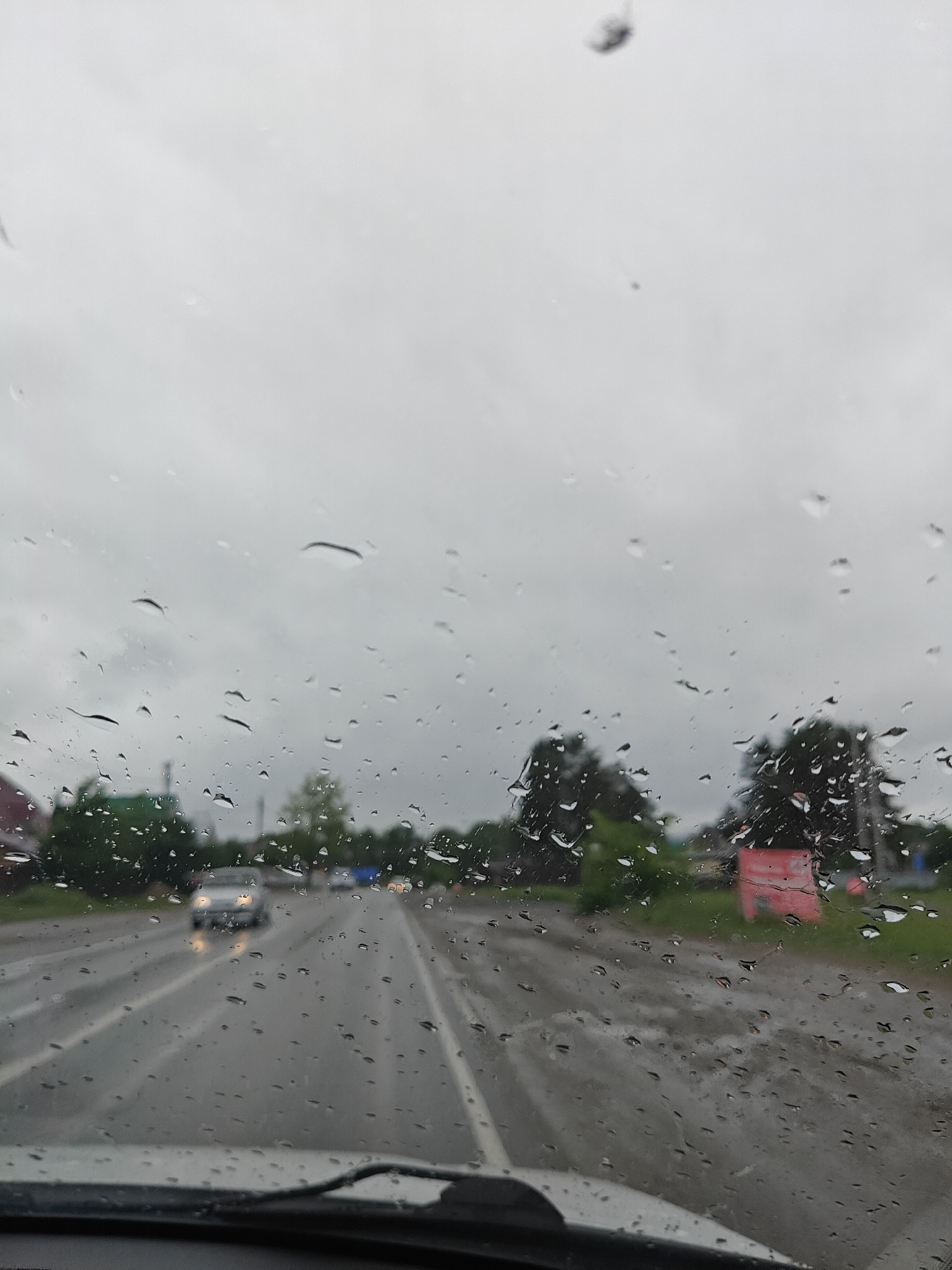The image appears to be taken from inside a vehicle, looking out through a rain-streaked windshield at a road and surrounding area. It is daytime, and the sky is overcast and grey, suggesting a rainy day. Several raindrops are visible on the windshield. A car with headlights on is traveling in the opposite direction on the left side of the road. There are other vehicles in the distance. Trees and some buildings, potentially houses or commercial structures, can be seen along the roadside. The road surface is wet, reflecting the ambient light. Based on the prompt stating the location is Chaikovsky, Russia, the vegetation and built environment are consistent with that region.
