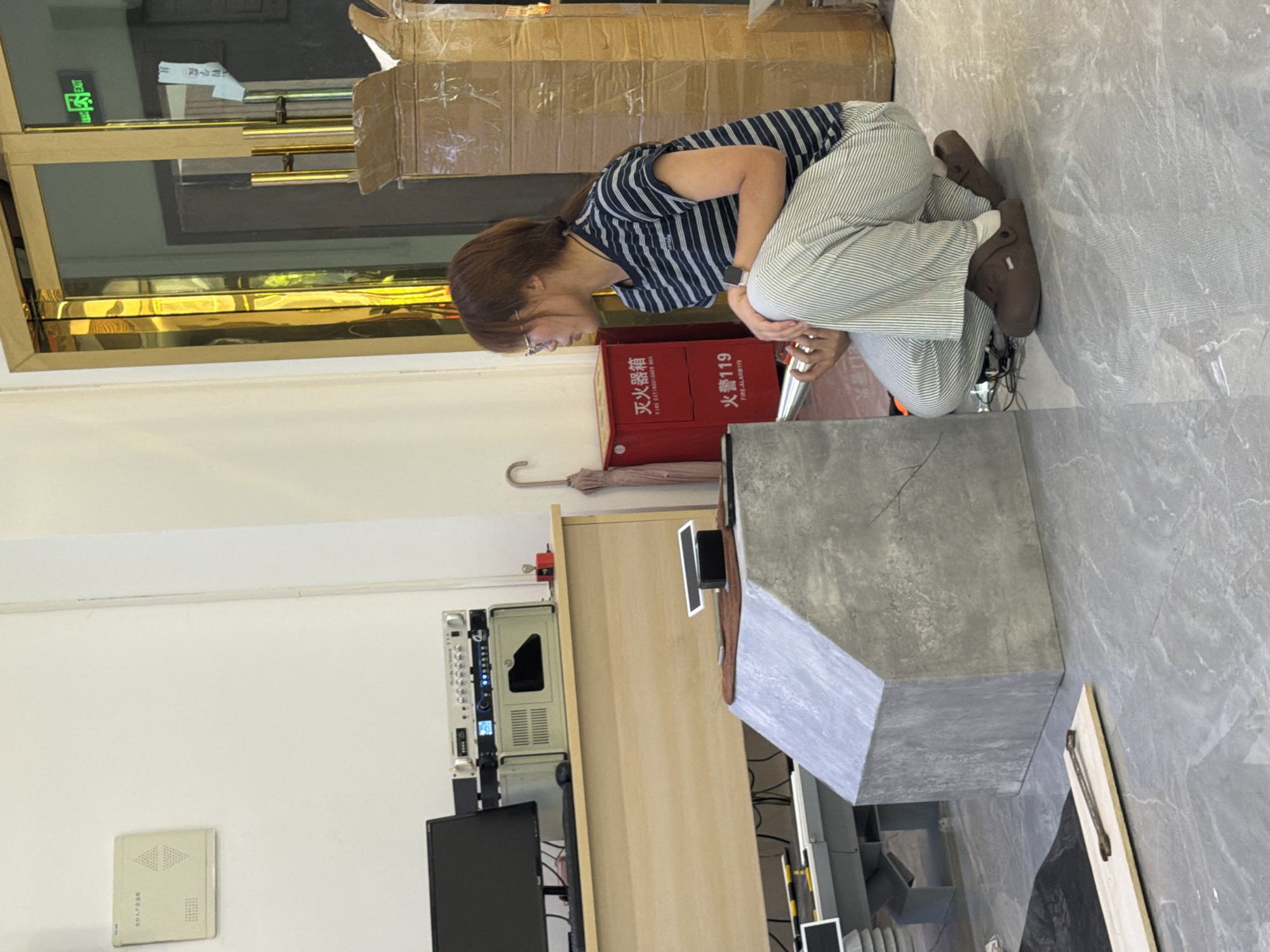In a brightly lit room, a young woman with brown hair tied back in a ponytail, wearing glasses, a navy and white striped t-shirt, and striped pants, squats on a grey tiled floor. She is holding a silver cylindrical object in her right hand and appears to be examining a concrete structure in front of her. To her left, a red fire extinguisher box is visible, with Chinese characters indicating "FIRE EXTINGUISHER BOX" and "FIRE ALARM 119". Behind her, a large cardboard box is stacked, and a light-colored wall features a white rectangular panel with Chinese writing. The room also contains a wooden desk with a computer monitor and audio equipment. An umbrella leans against the wall near the fire extinguisher. The scene appears to be an interior commercial space, possibly a workshop or retail establishment, located in Jiangjin, China, given the presence of Chinese signage and the overall ambiance.
