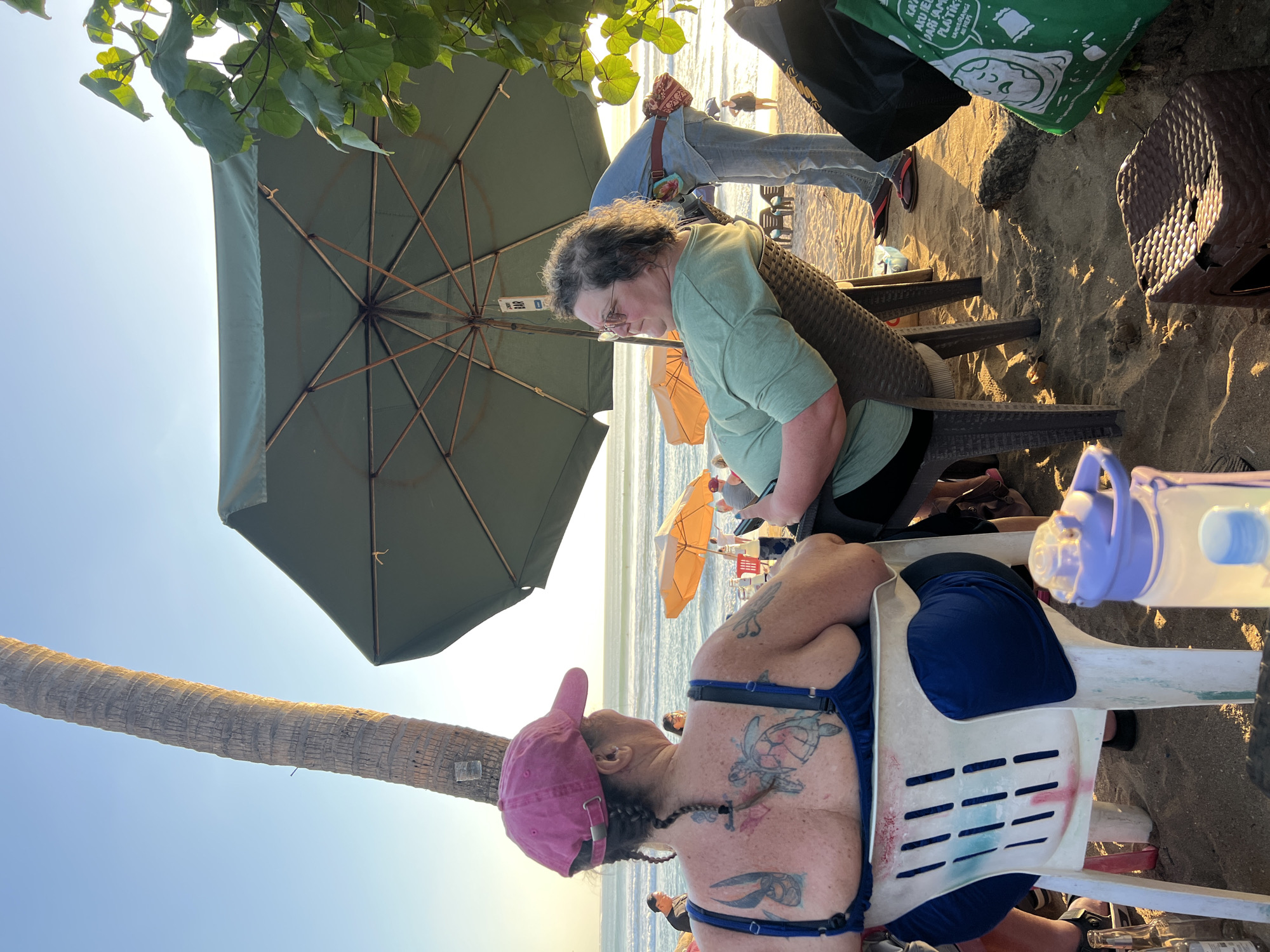 Two women are seated at tables on a sandy beach in Kuta, Indonesia, overlooking the ocean. A large green umbrella provides shade overhead. The woman in the foreground, viewed from behind, wears a pink baseball cap and a dark blue swimsuit. Her back is adorned with tattoos, including a turtle and a butterfly. She is seated in a white plastic chair. To her right, another woman wearing glasses and a light green t-shirt is seated in a dark brown wicker chair, facing the ocean. In the background, several other people are visible, along with orange beach umbrellas and lounge chairs. A tall palm tree stands to the left of the frame. The setting sun casts a warm glow on the sand and water, indicating it is late afternoon or early evening.