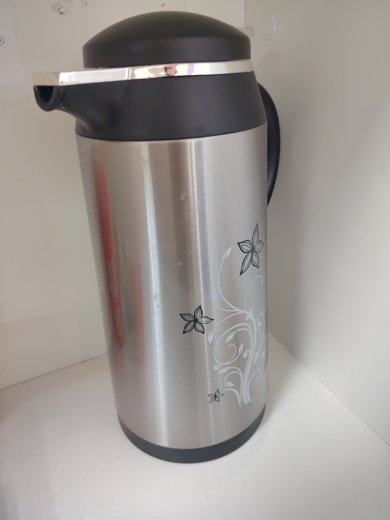 A tall, silver thermos with a black lid and handle sits on a white surface. The thermos has a floral design on its side. The background is a plain white wall.