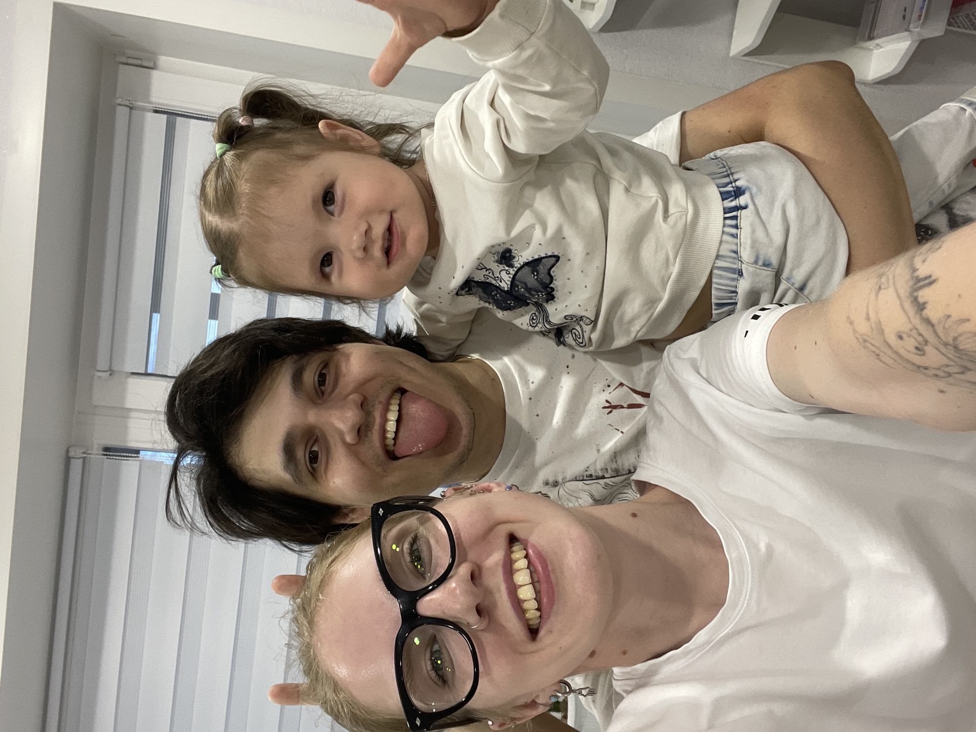 Three people are posing for a selfie indoors. A fair-skinned woman with blonde hair and black-framed glasses is in the foreground, smiling with visible teeth. Behind her and slightly to the left is a man with dark hair who is sticking out his tongue. At the top of the frame, a toddler with fair skin, light brown hair tied in pigtails, is reaching up with one hand. All three individuals are wearing white shirts. The background includes white blinds on a window and a white shelf. There is no indication of the location being Izhevsk, Russia.