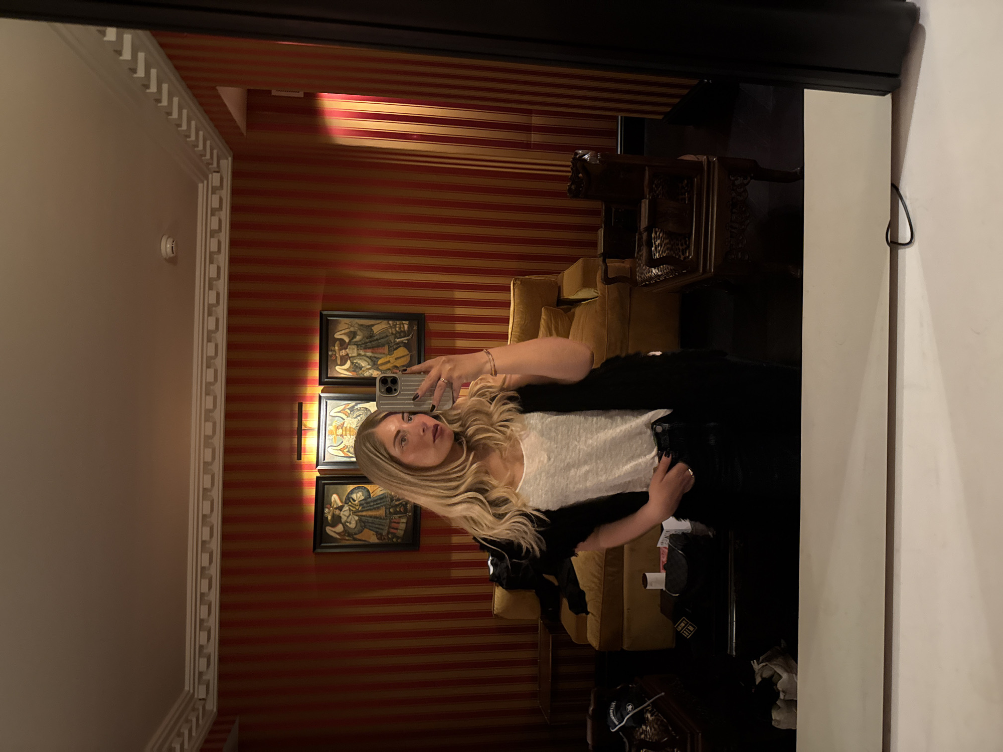 A woman is taking a selfie in a mirror. She has long blonde hair and is wearing a white t-shirt and a black jacket. The room is decorated with red and gold striped wallpaper and three framed paintings. There is a yellow couch and a dark wood table in the background. It is not possible to determine the location of this room within the city of Paris, France with only visual information.
